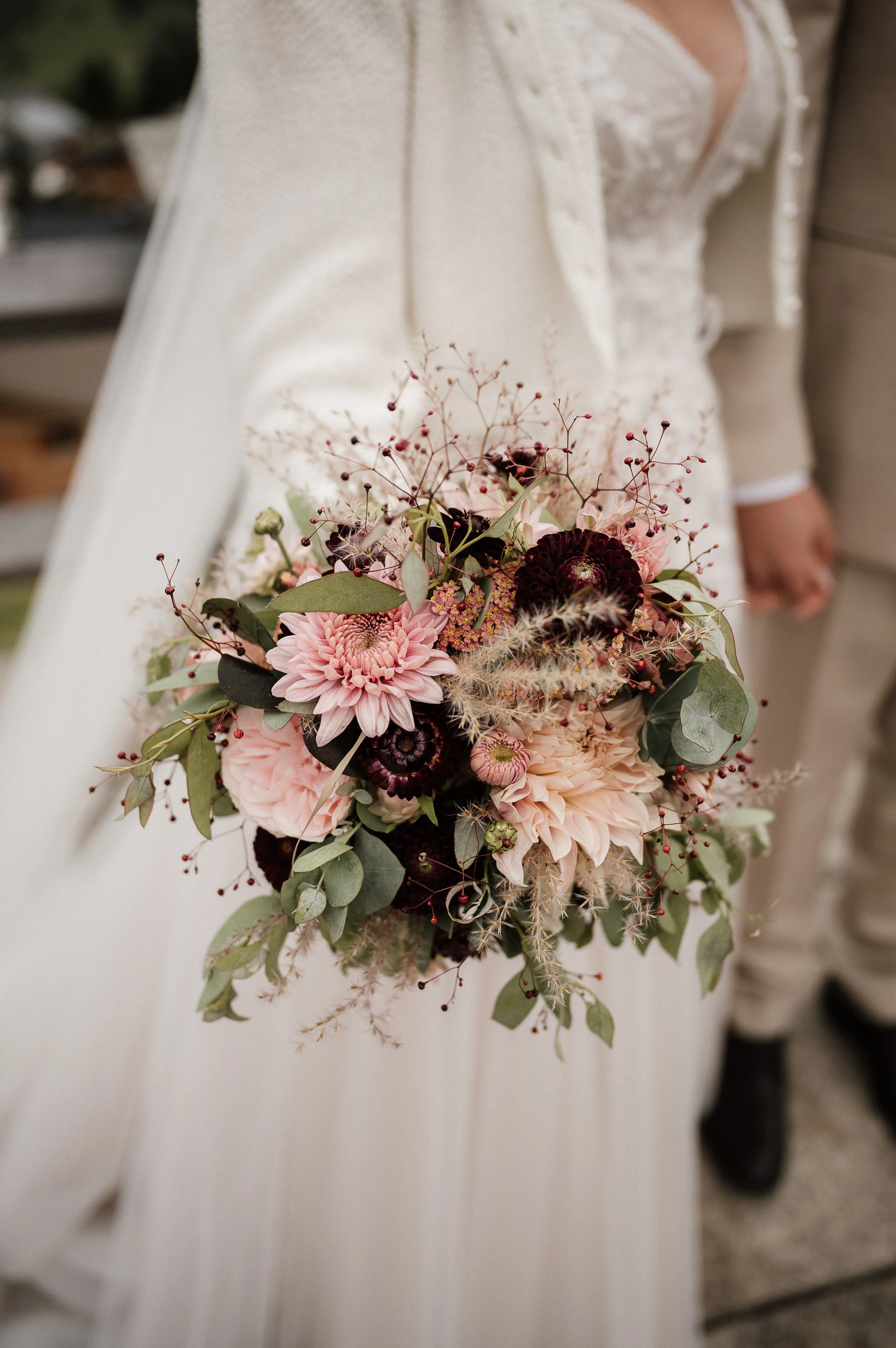 Braut hält einen Blumenstrauß mit pinken, weißen und dunklen Blumen sowie grünem Laub bei einer Hochzeit.