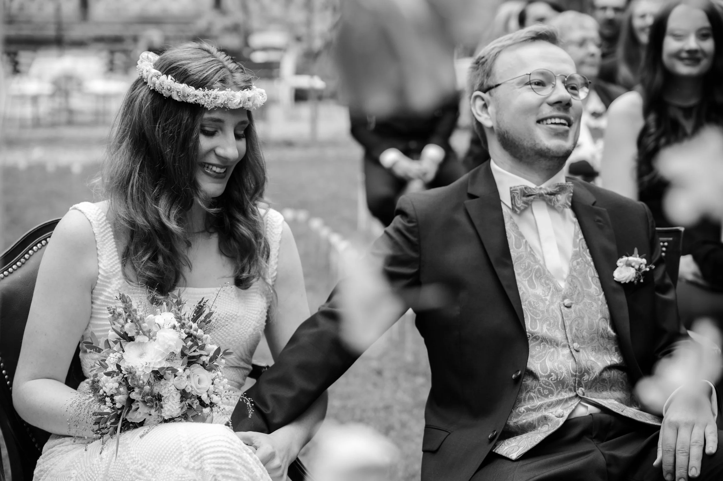 Ein Brautpaar bei einer Hochzeitszeremonie, beide lächeln glücklich, die Braut trägt eine Blumenkrone und hält einen Blumenstrauß, der Bräutigam trägt eine Anzugjacke mit Weste und Fliege. Es sind weitere Gäste im Hintergrund.