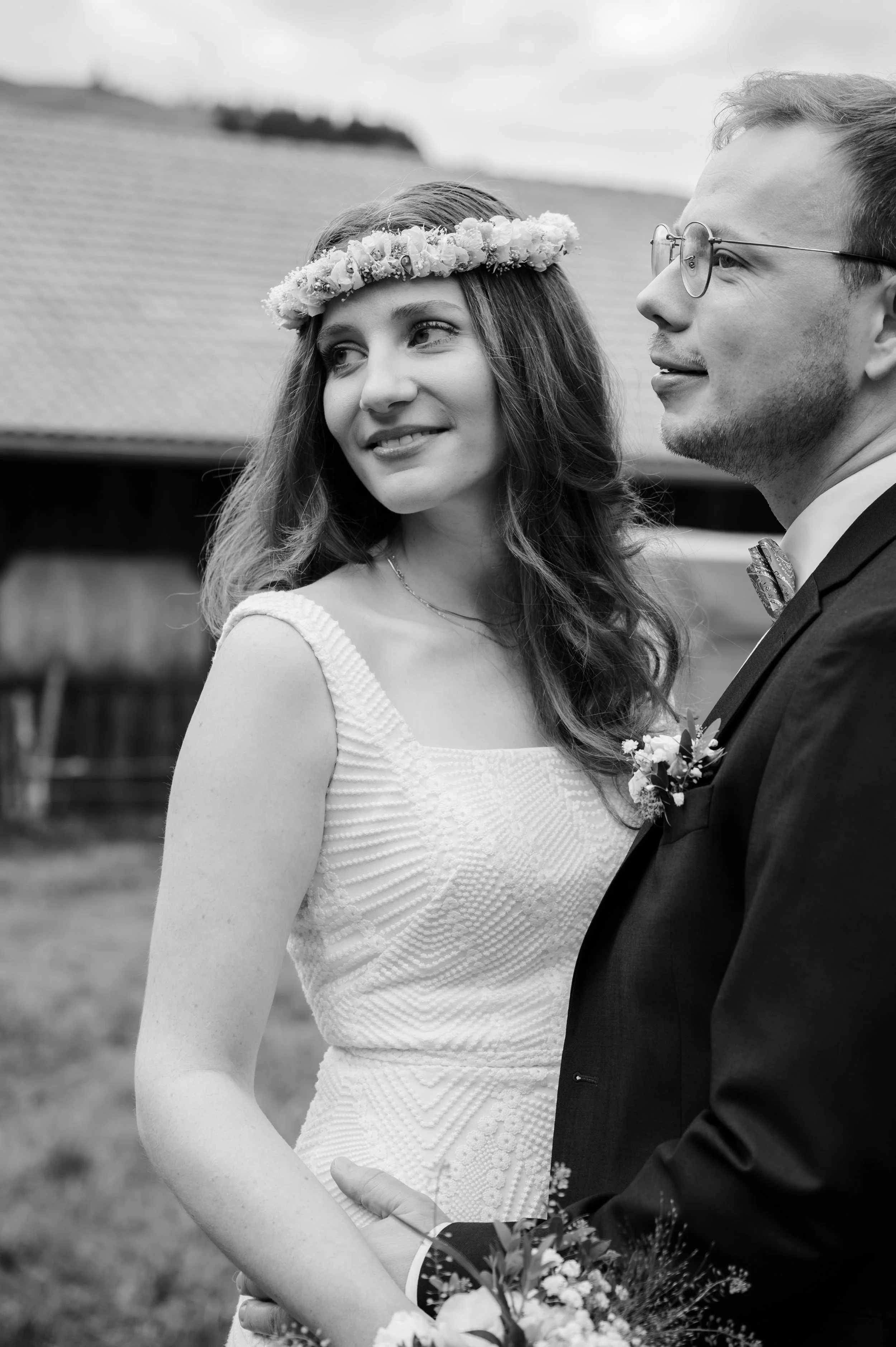 Ein Brautpaar bei einer Hochzeit im Freien, die Braut trägt ein weißes Kleid und einen Blumenkranz im Haar, der Bräutigam trägt einen Anzug mit Fliege, beide schauen sich liebevoll an.