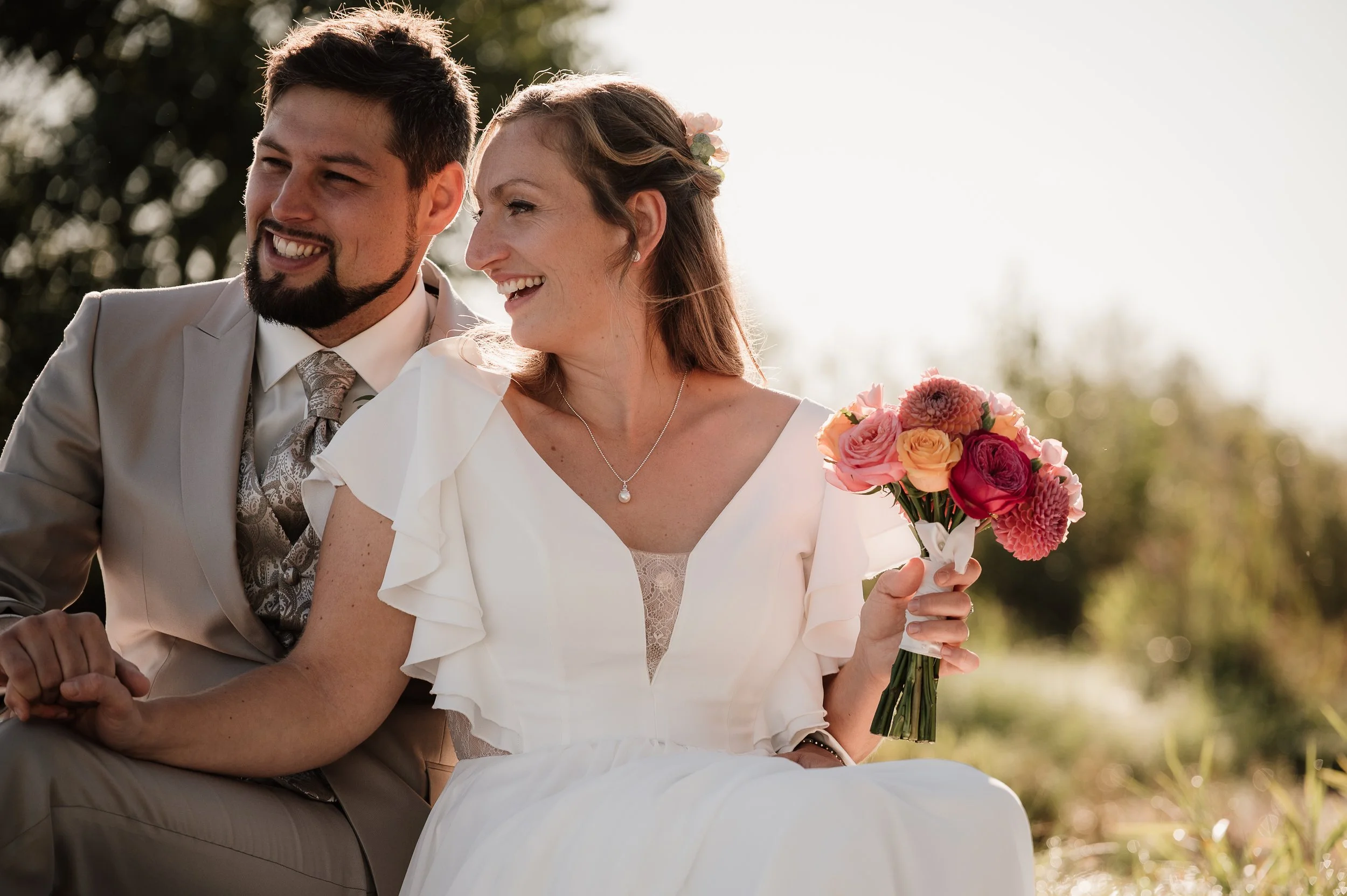 Ein frisch verheiratetes Paar sitzt im Freien, die Frau hält einen Blumenstrauß und beide lächeln glücklich.