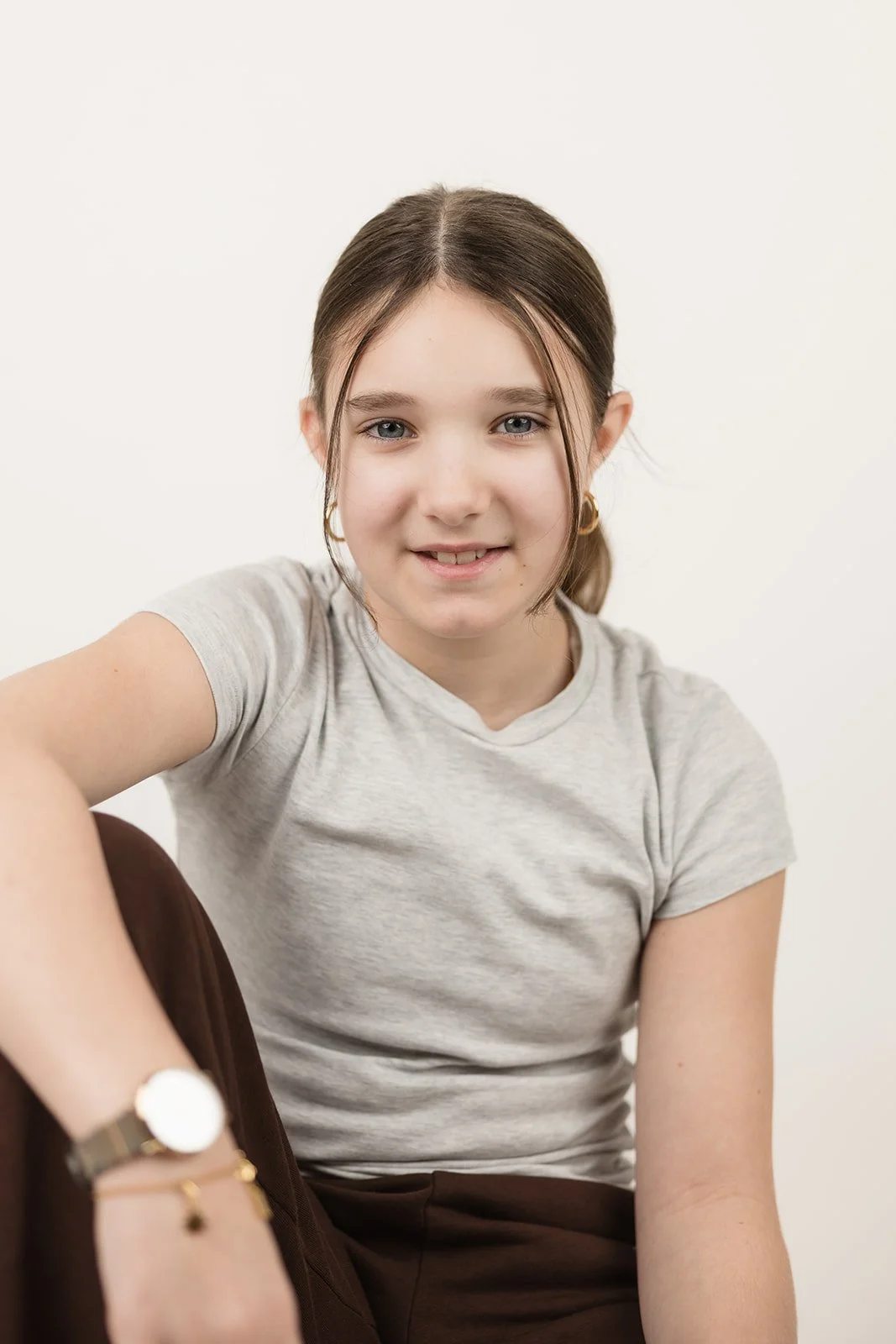 Nahaufnahme eines jungen Mädchens mit braunen, glattgesteckten Haaren, blauen Augen und Ohrringen, sitzend vor weißem Hintergrund, casual Kleidung, freundliches Lächeln.