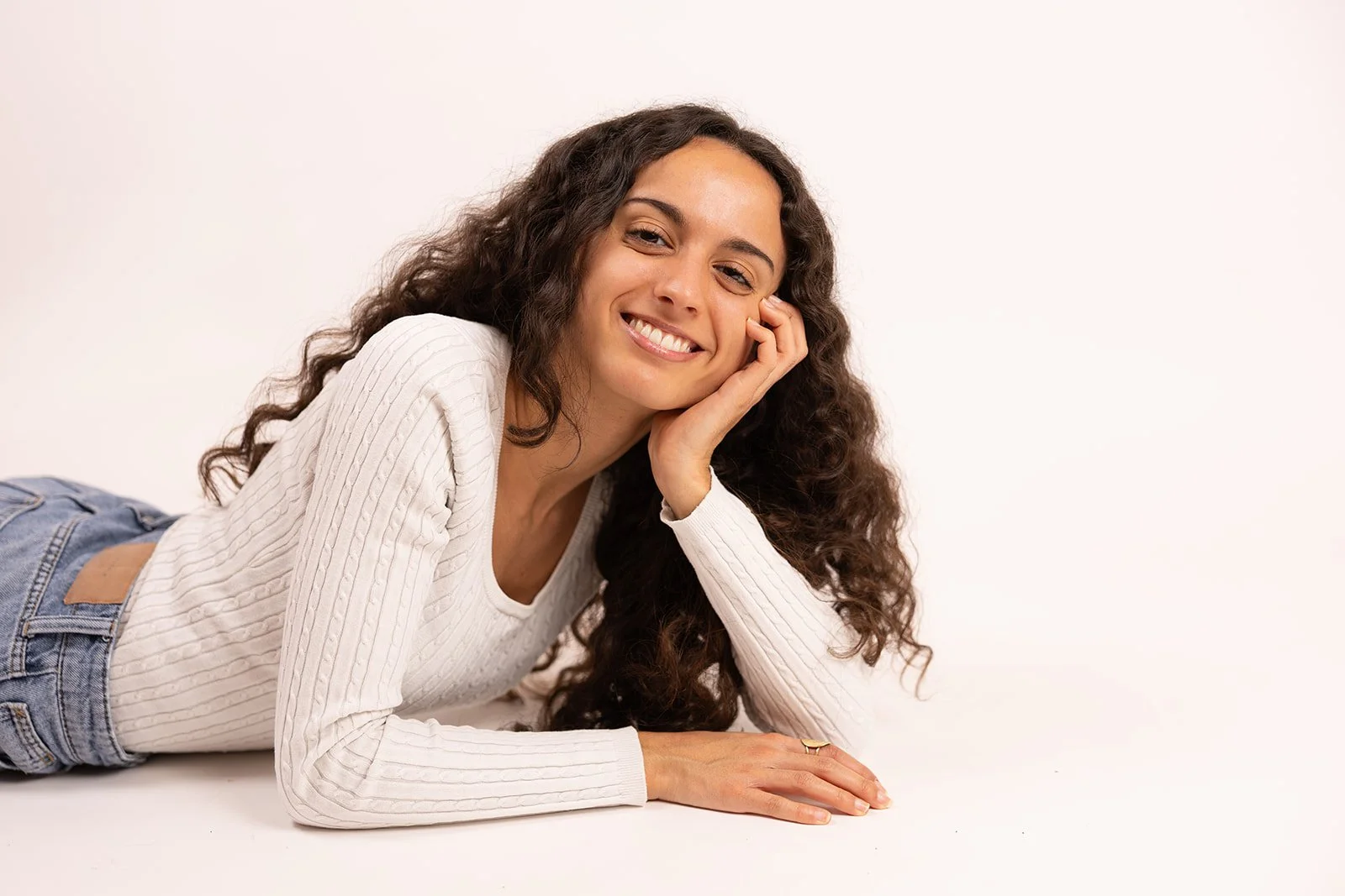 Lächelnde junge Frau mit lockigem Haar liegt auf dem Bauch auf weißem Hintergrund, trägt weiße Strickjacke und Jeans.