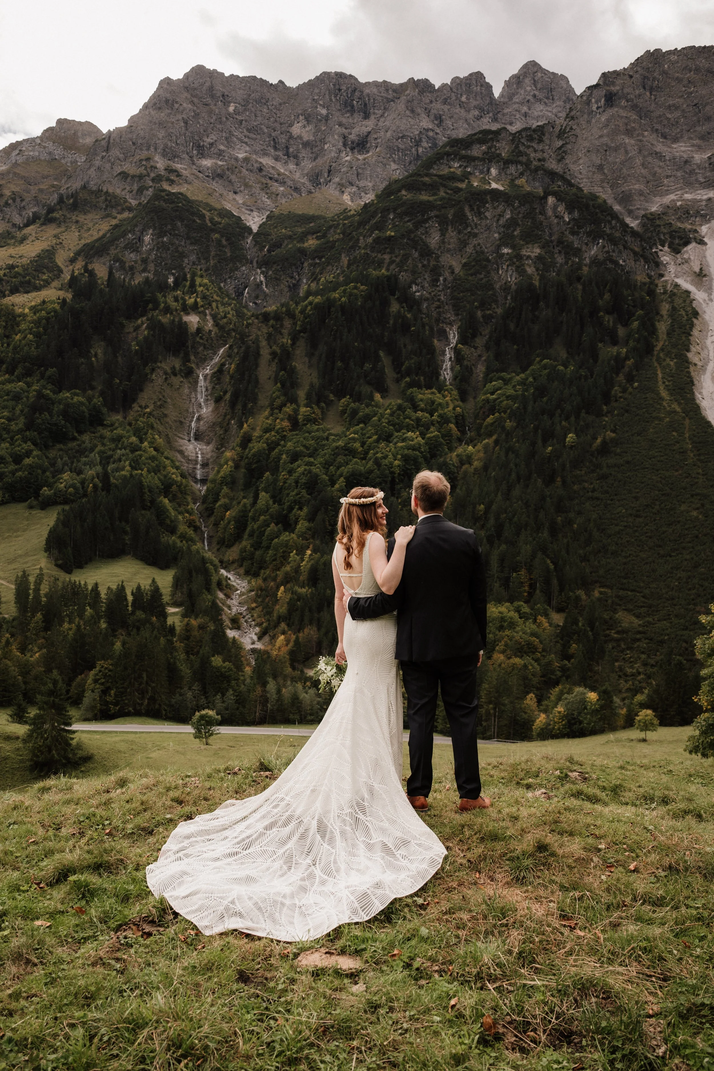 Braut und Bräutigam stehen auf einer Wiese in den Bergen, mit Blick auf einen bewaldeten Hügel und Wasserfälle im Hintergrund.