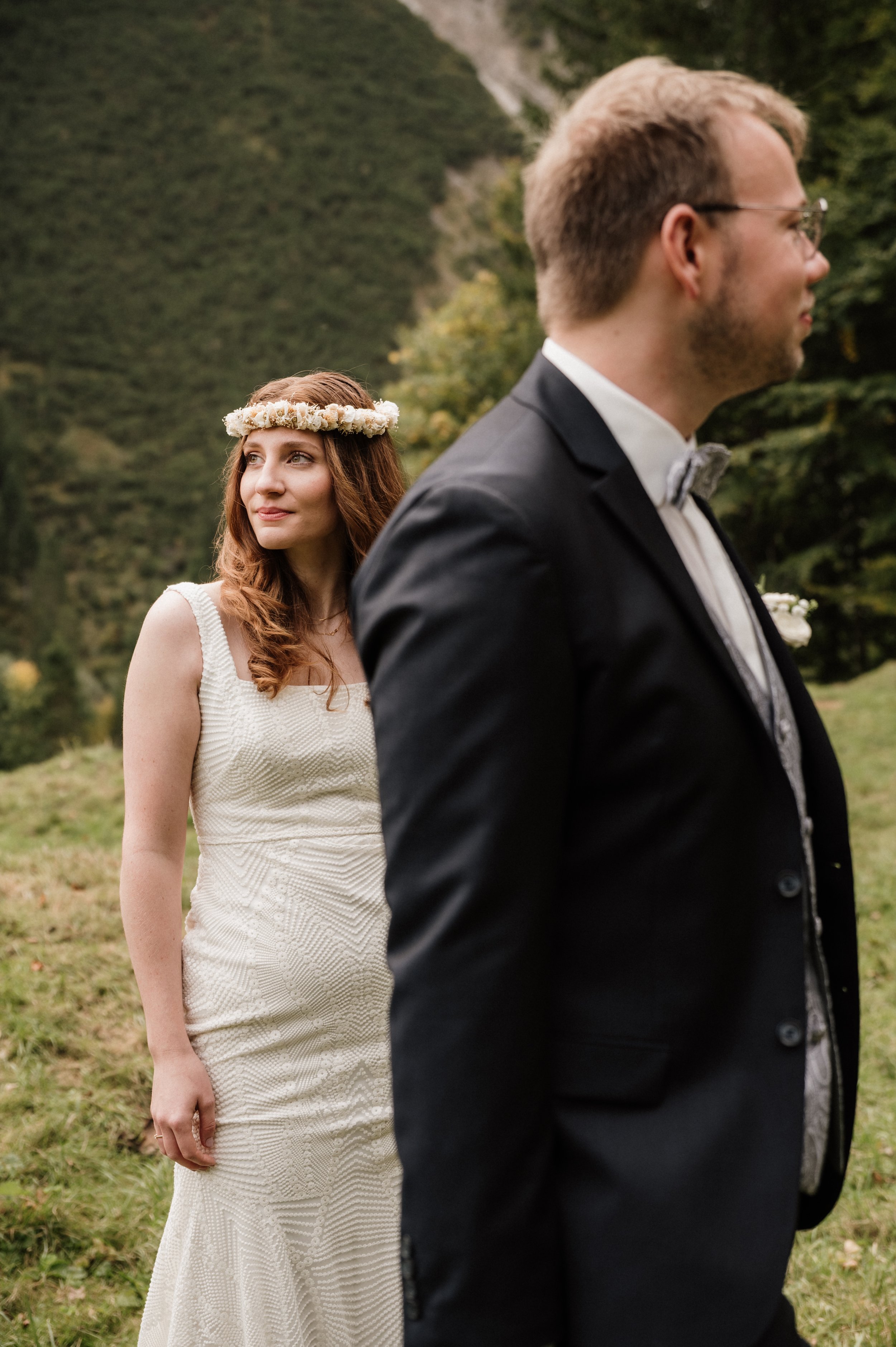 Ein junges Paar bei einer Hochzeit im Freien, die Braut trägt ein weißes Hochzeitskleid mit einer Blumenkrone im Haar, während der Bräutigam einen schwarzen Anzug trägt, und sie stehen im Grünen in einer malerischen Landschaft.