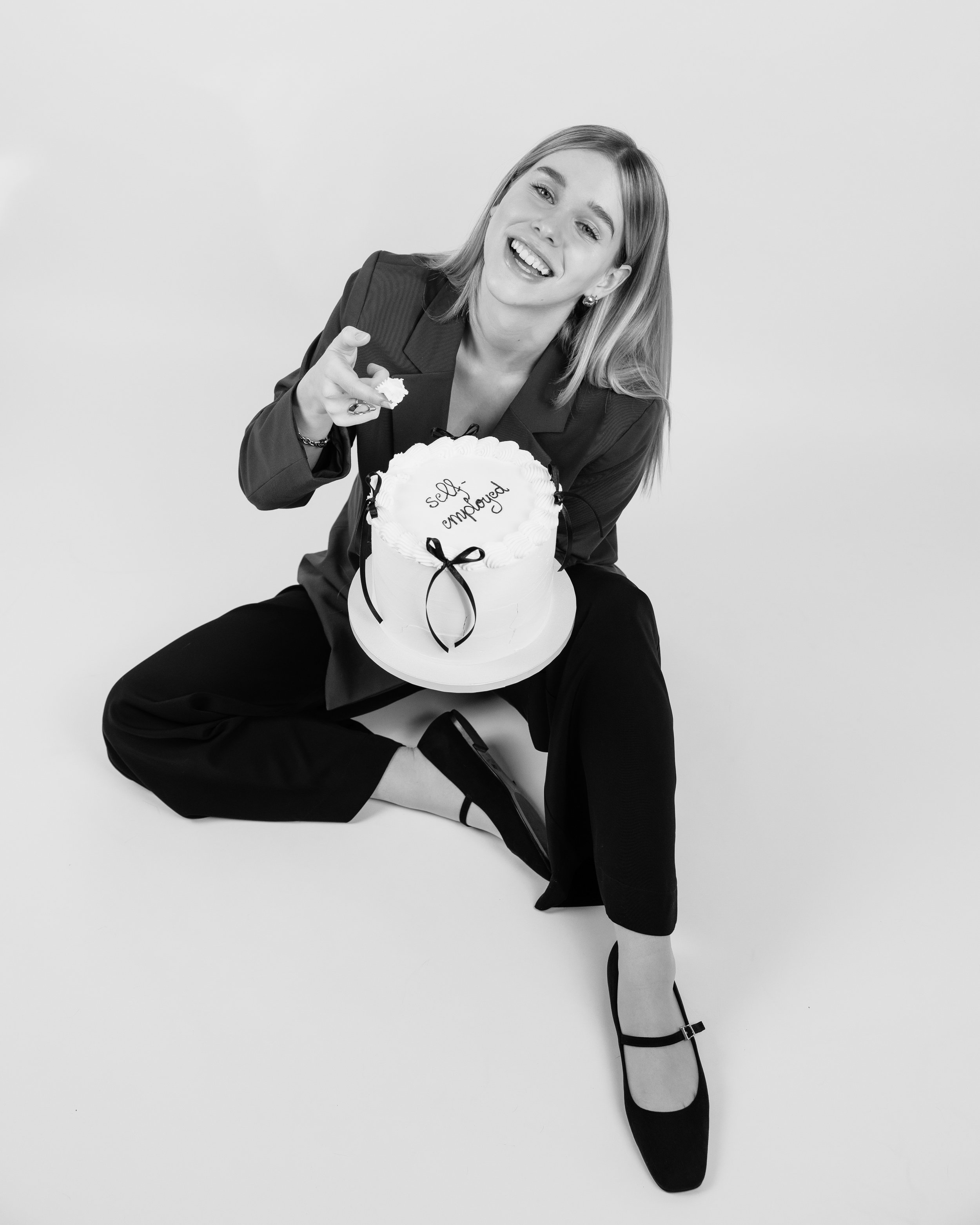 Schwarz-weiße Fotografie einer jubelnden Frau in Businesskleidung mit einem 'Self Employed' Kuchen.