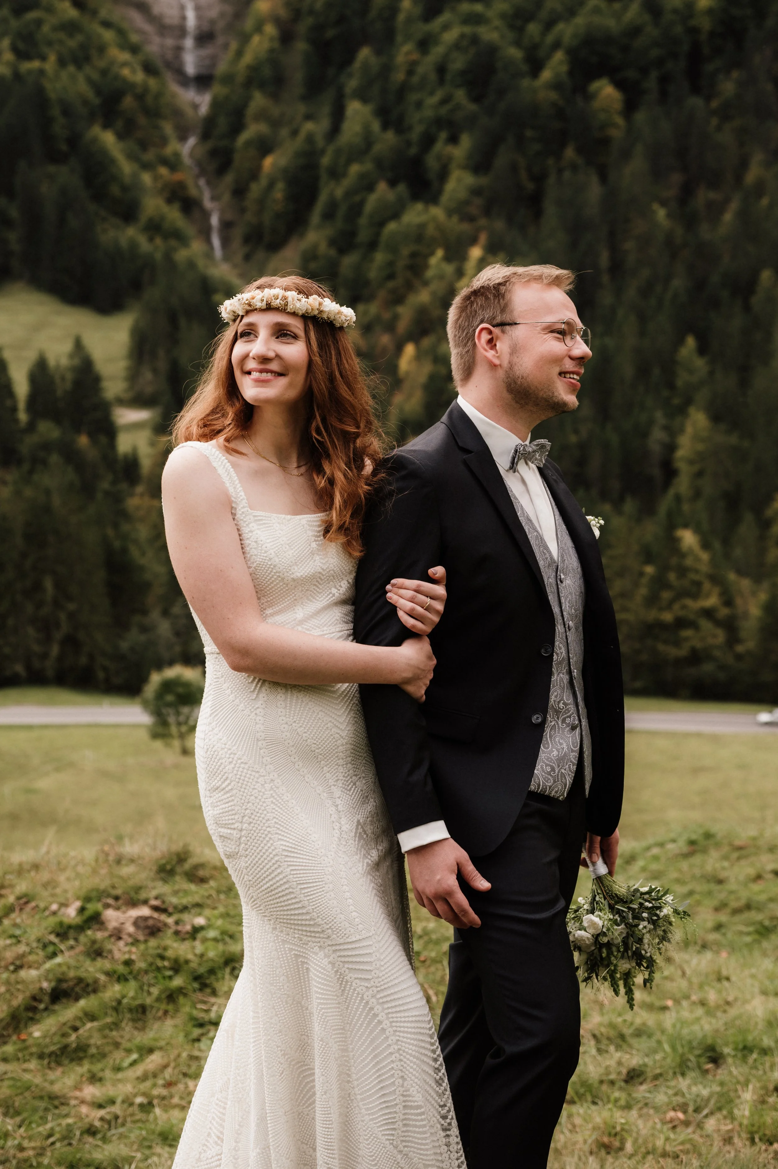 Ein Brautpaar steht im Grünen, die Braut trägt ein langes, weißes Hochzeitskleid und einen Blumenkranz im Haar, während der Bräutigam einen schwarzen Anzug und eine Fliege trägt, in der Hand hält er einen Blumenstrauß, im Hintergrund sind Bäume und ein Wasserfall an einem bewölkten Tag.