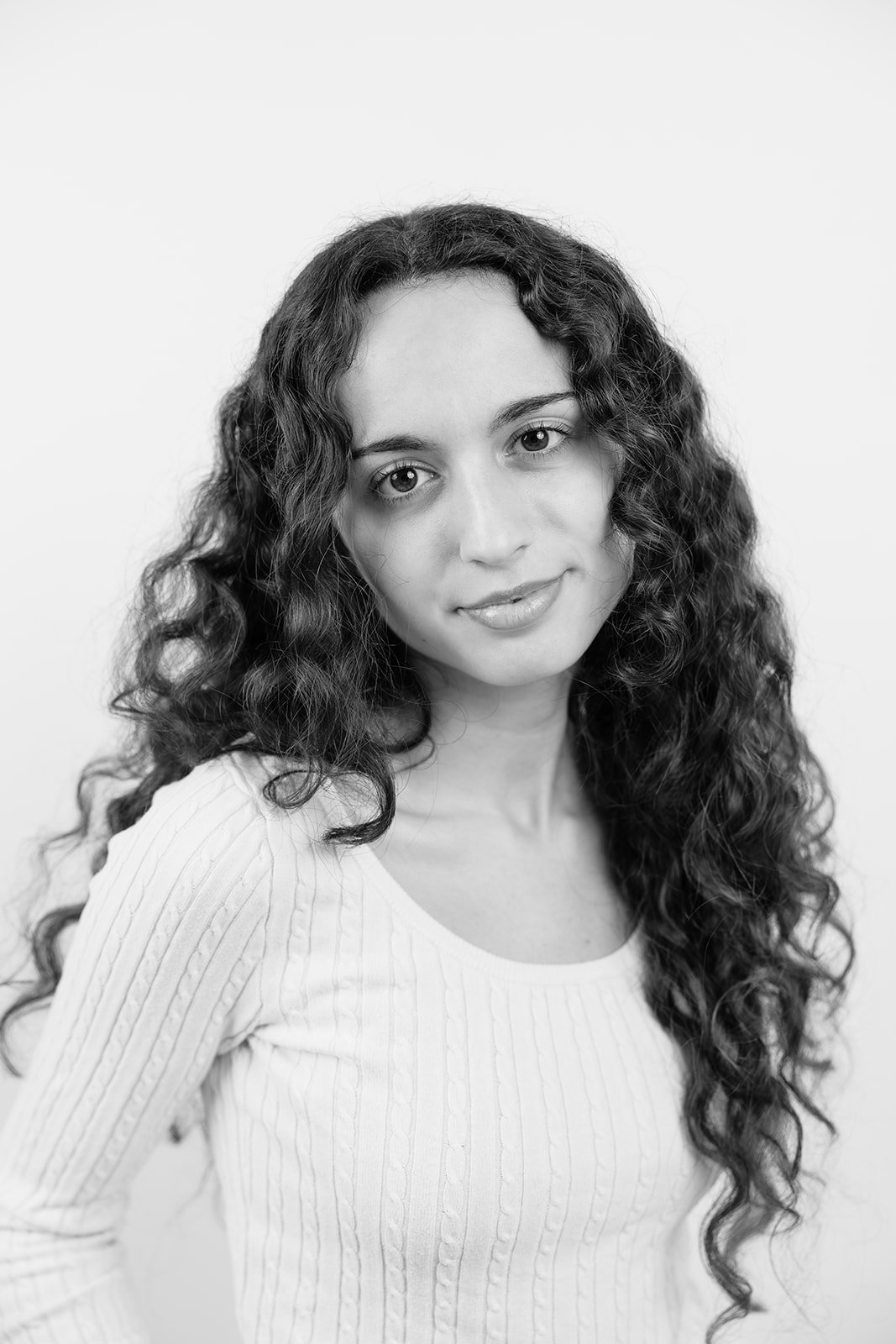 Schwarz-Weiß-Porträt einer jungen Frau mit langen, lockigen Haaren, die eine helle, gerippte Bluse trägt, vor hellem Hintergrund.