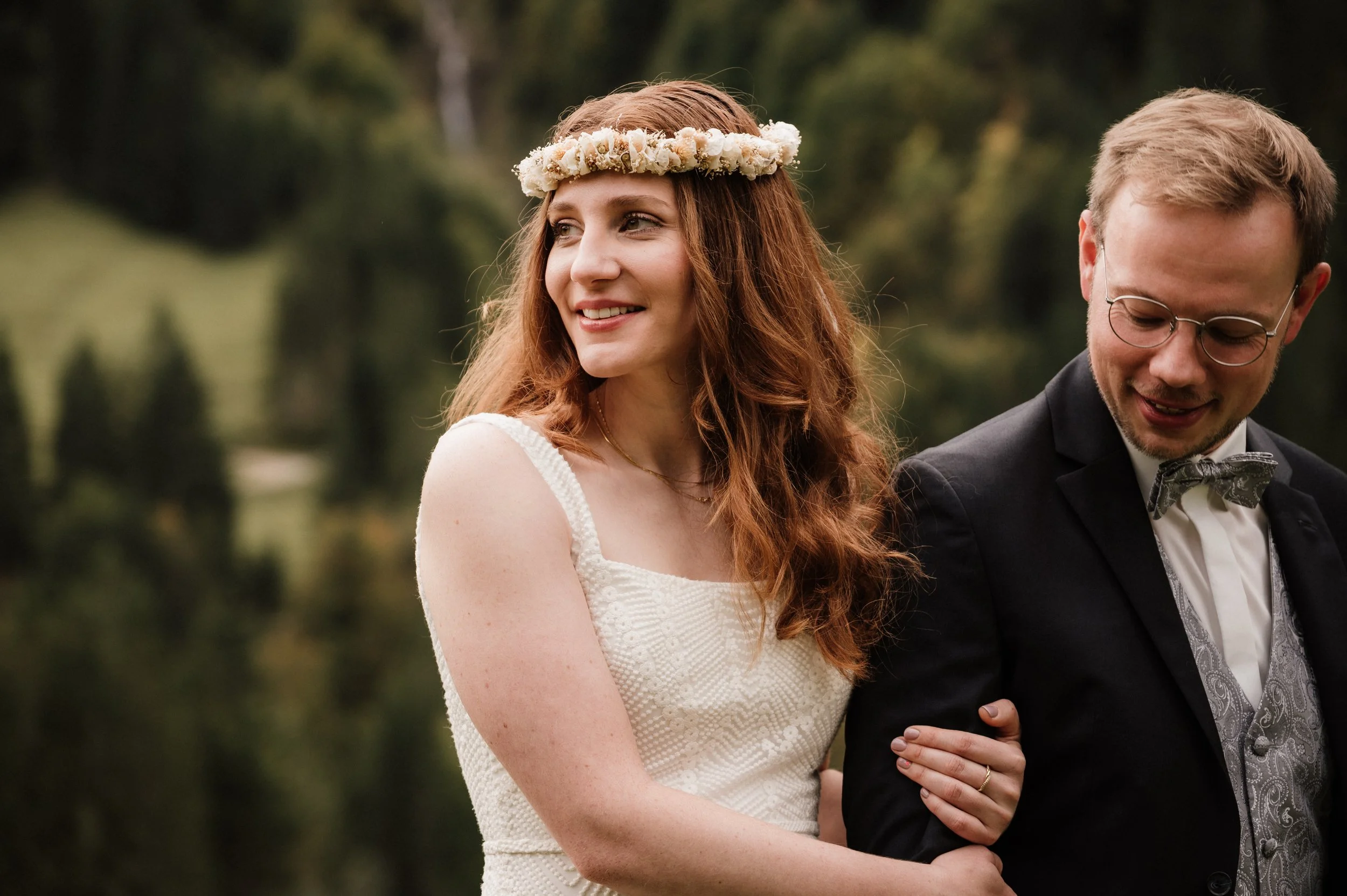 Braut mit lockigem rotbraunem Haar trägt Blumenkronen-Haarband und weißes Kleid, während Bräutigam mit Brille und Anzug lächelt, bei einer Hochzeit im Freien.