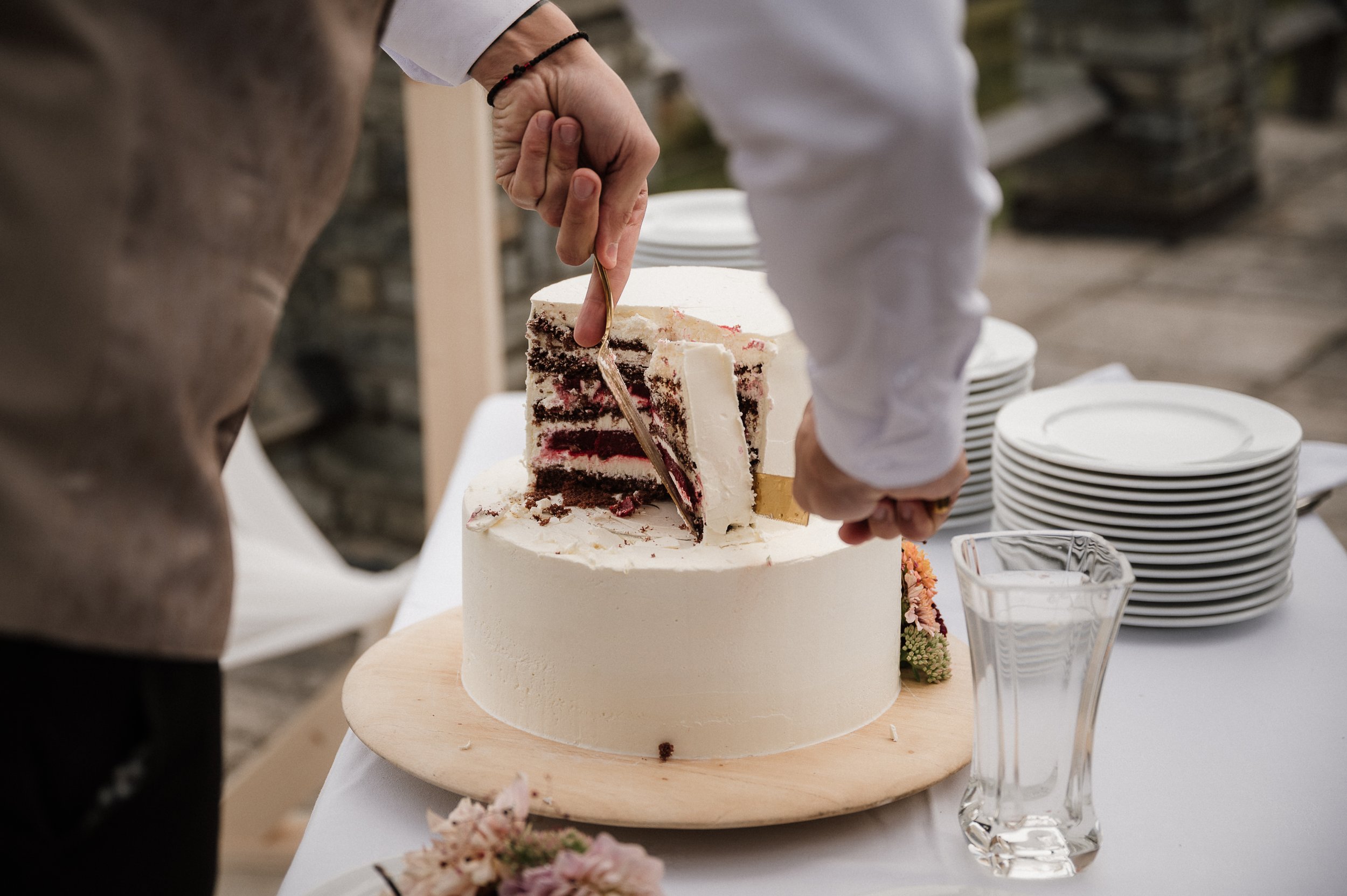 Person schneidet eine mehrstöckige Hochzeitstorte mit einem Messer. Die Torte ist weiß und hat eine Füllung aus roten Beeren. Es gibt weiße Teller und ein leeres Wasserglas auf dem Tisch.