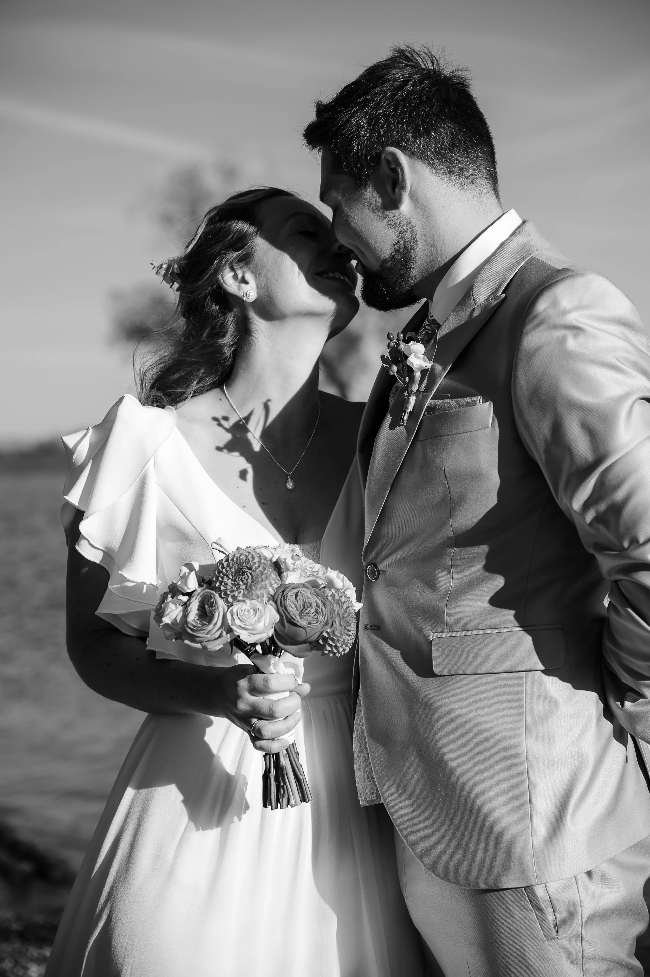 Ein schwarzer und weißer Hochzeitsfoto von einem Brautpaar, das sich liebevoll berührt, mit Blicken, die sich fast küssen, draußen mit Himmel und Wasser im Hintergrund, die Braut hält einen Blumenstrauß.