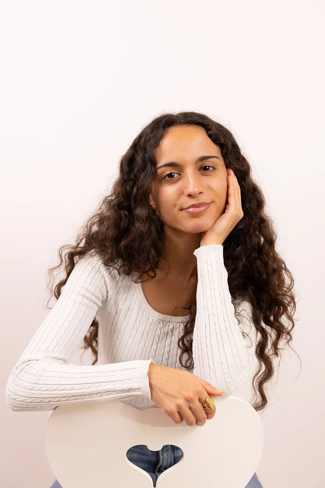 Junge Frau mit langen, lockigen braunen Haaren in weißem Pullover lehnt an einem weißen Stuhl mit Herzgesicht-Ausschnitt vor einem weißen Hintergrund.
