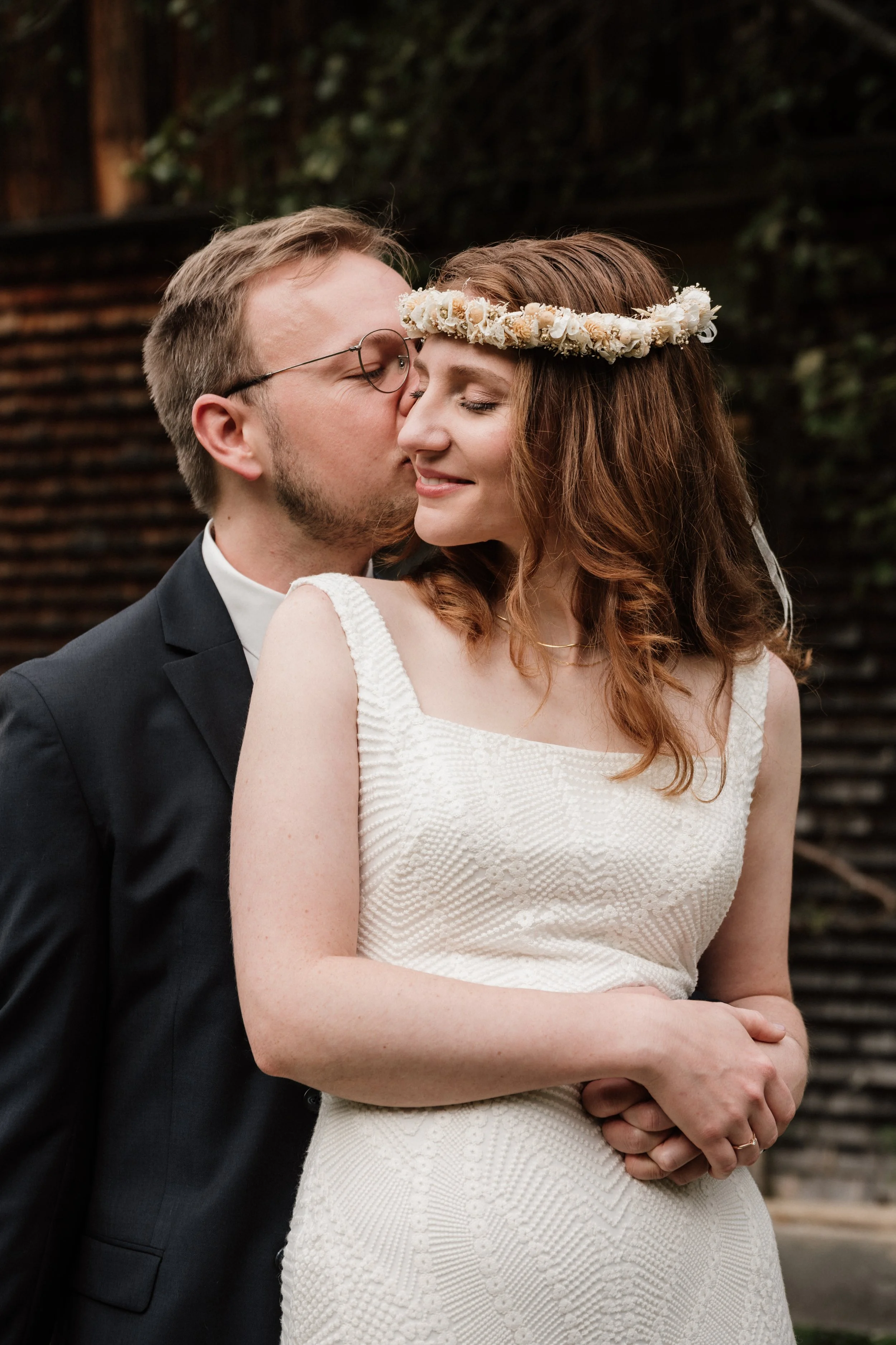 Ein Brautpaar beim Hochzeitsportrait, der Bräutigam küsst die Braut auf die Wange, die Braut trägt eine Blumenkronen und ein weißes Kleid, im Hintergrund ein Holzzaun und Grün.