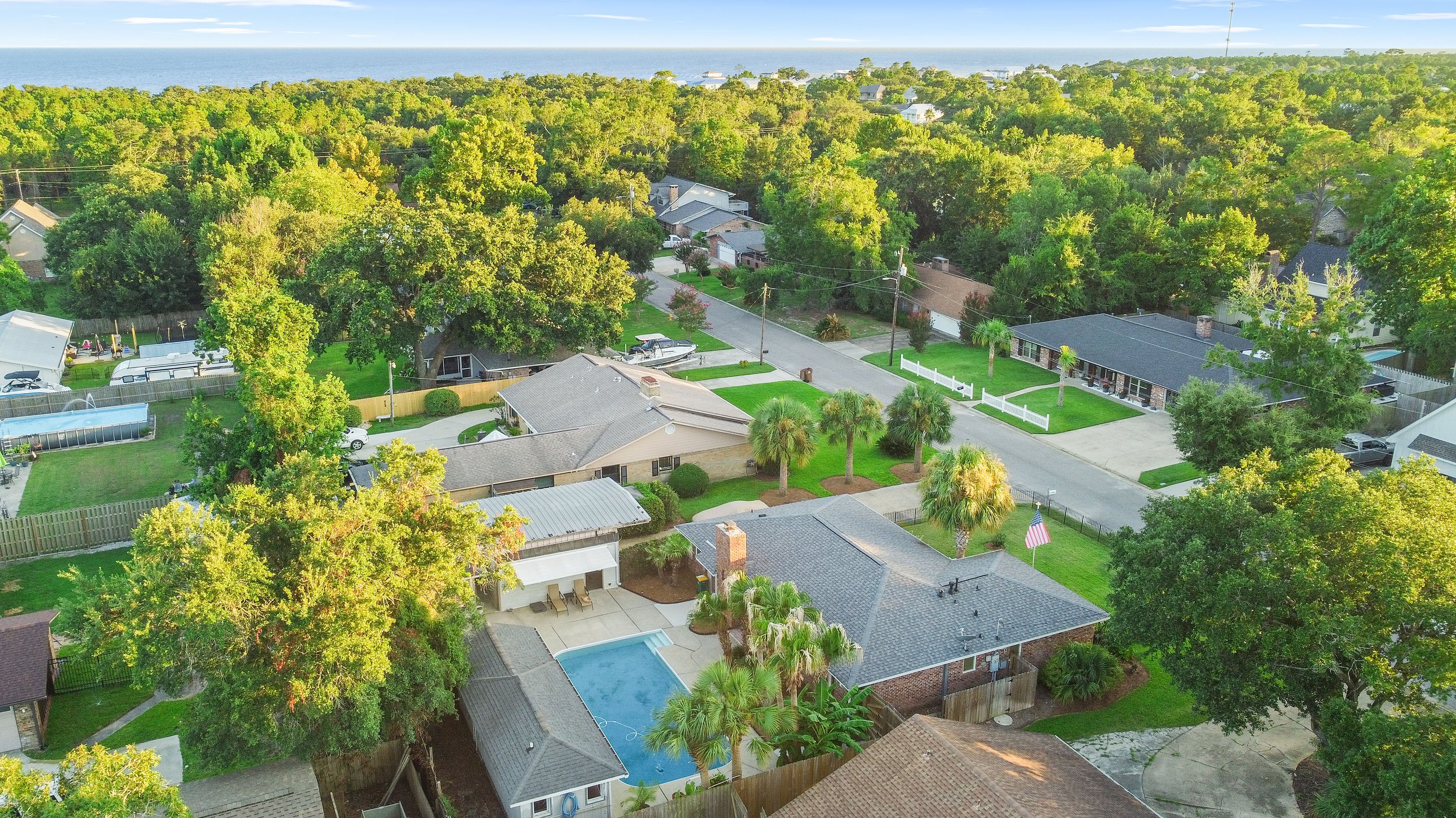 Aerial view of a suburban neighborhood with houses, trees, and a swimming pool. The ocean is visible in the background.