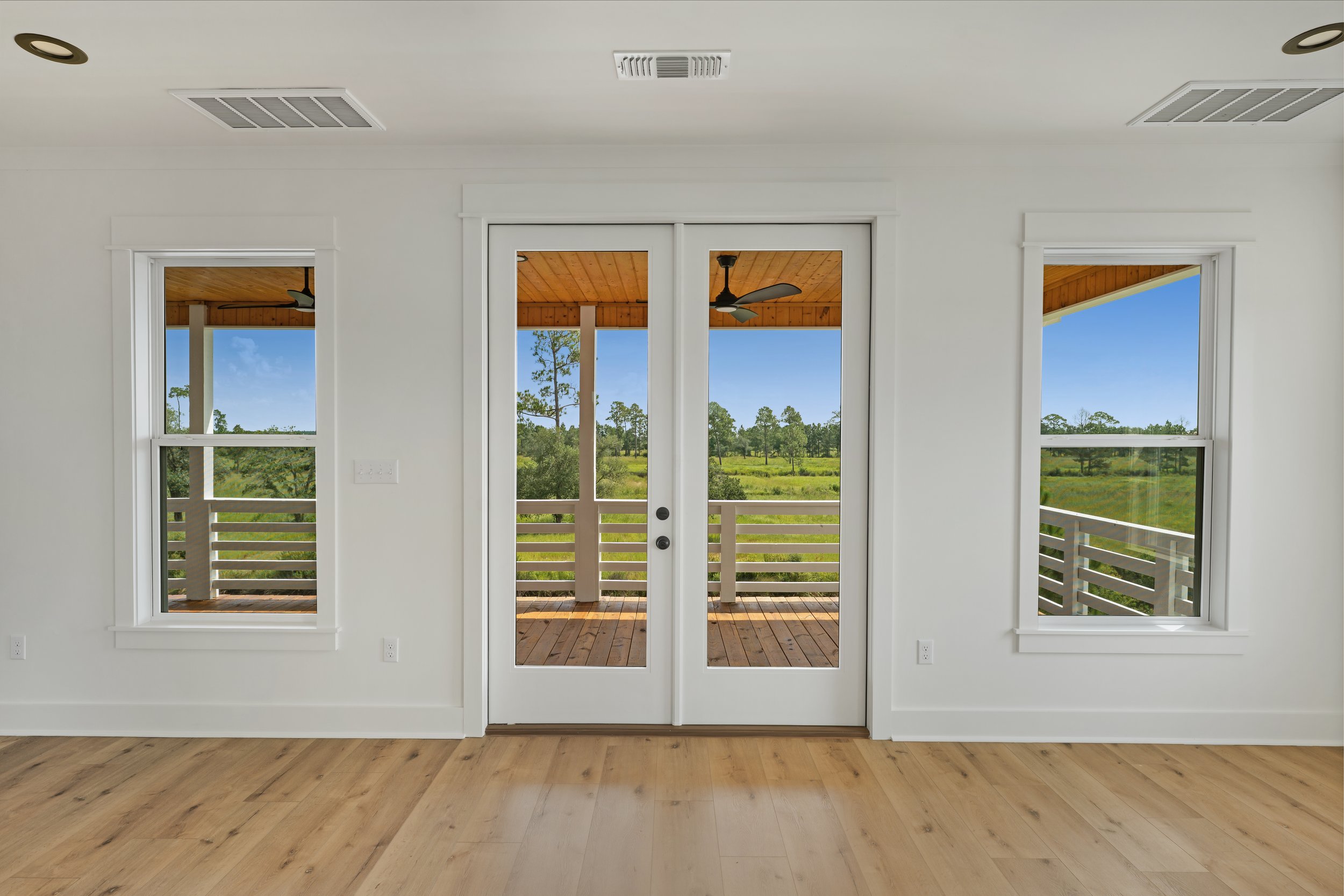 Interior view of a room with white walls, light wood floor, and large windows looking out onto a deck and green landscape with trees and blue sky.