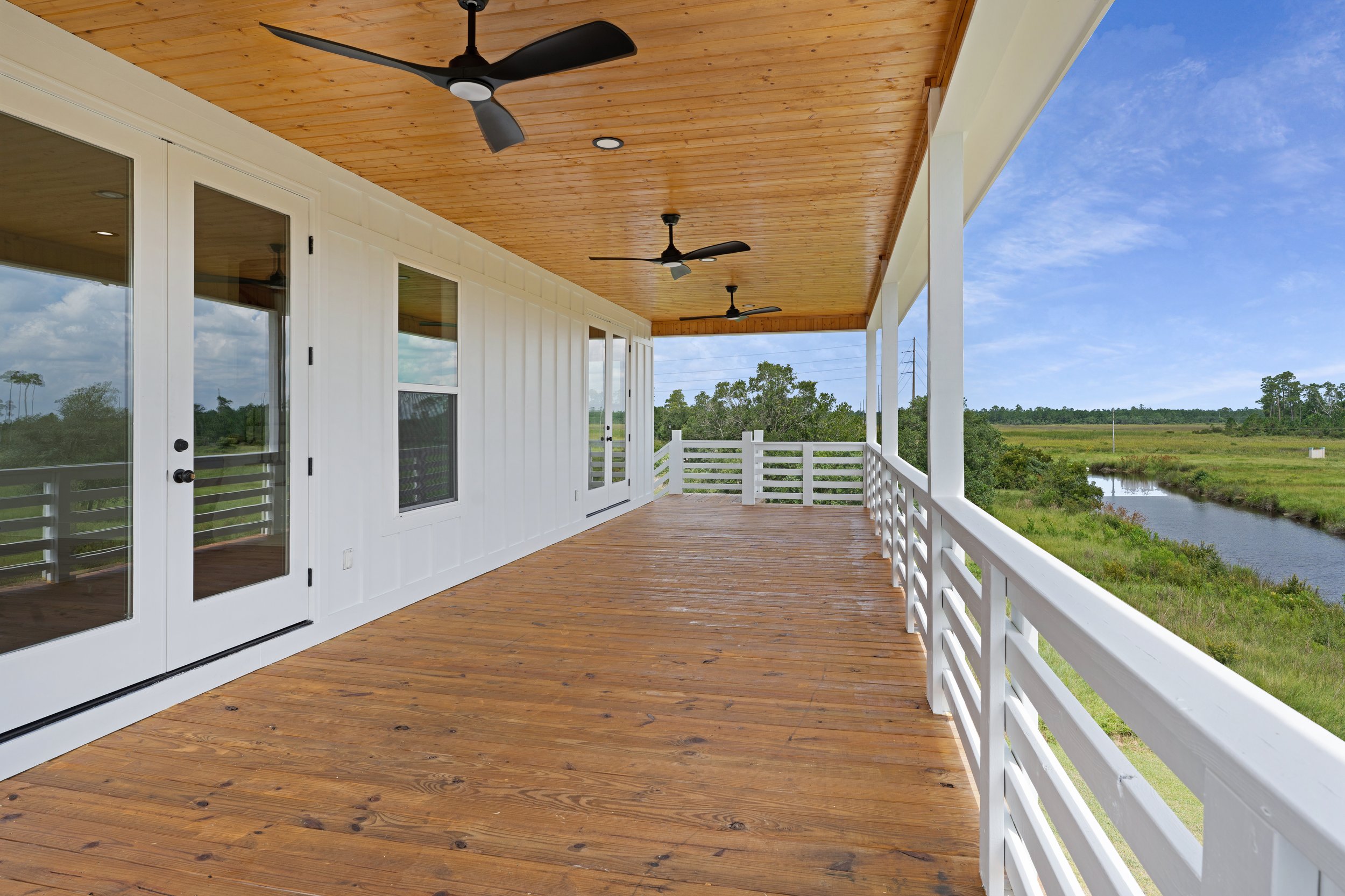 Empty front porch with wooden flooring, white railings, ceiling fans, and a view of a marshland with water and green trees in the distance.