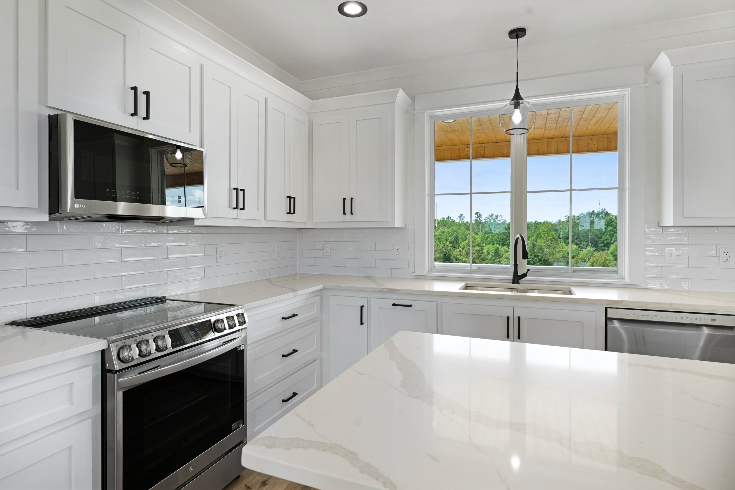 Modern white kitchen with large window showing green trees, stainless steel stove, microwave, dishwasher, white cabinetry, and a central island with a white marble countertop.