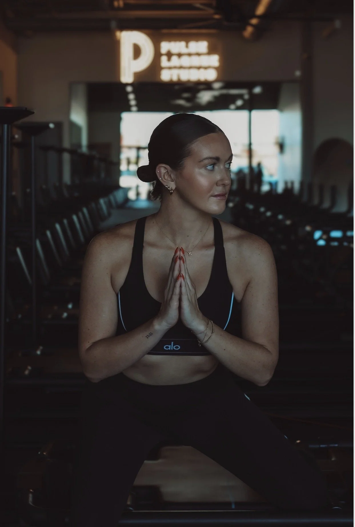 A woman in workout clothes sitting on a workout bench at the gym with her hands pressed together in a prayer position, looking to the right.