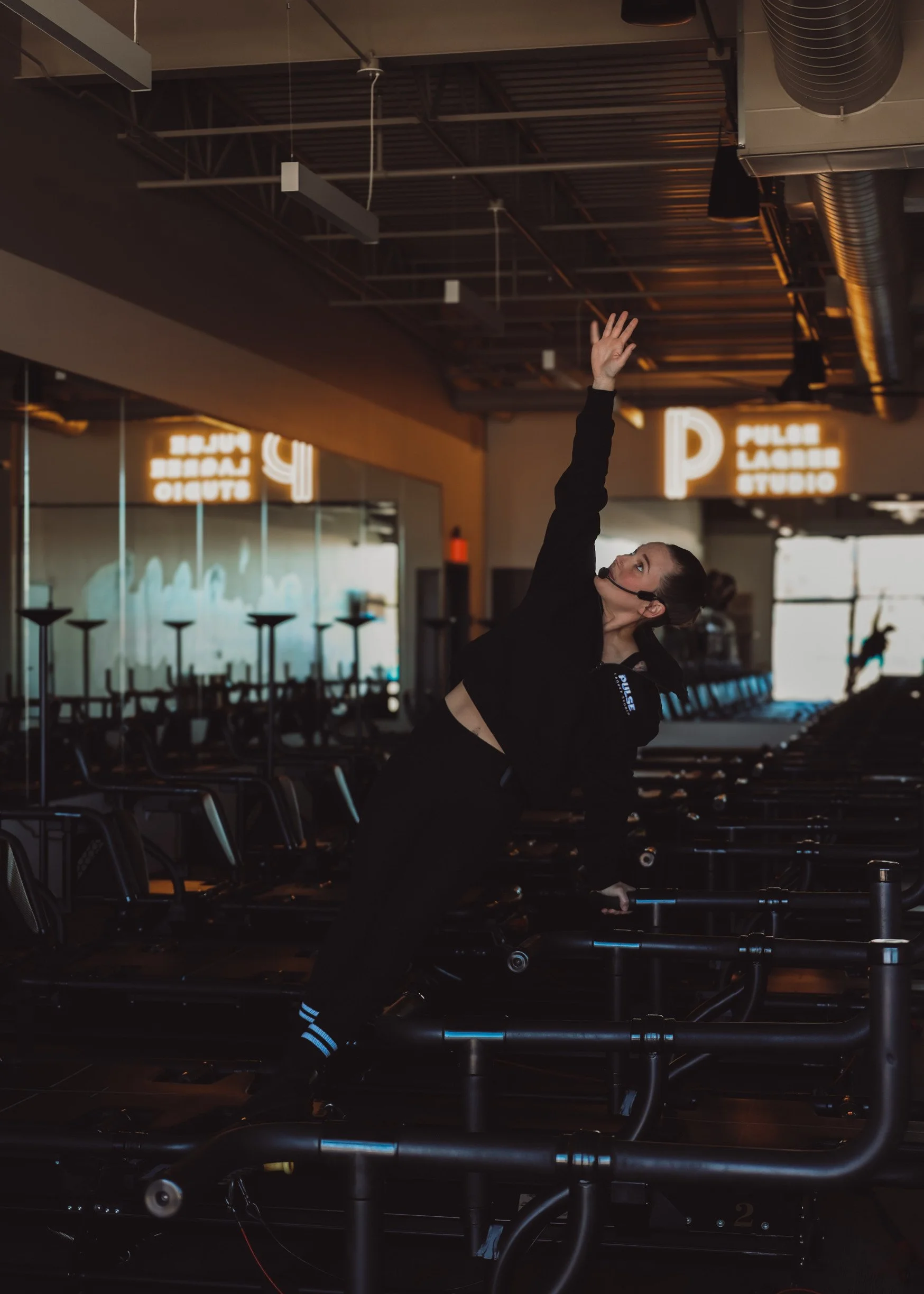 A woman in black athletic clothing and a headset stretches with her arm extended upward at a fitness studio, with exercise equipment and neon signage in the background.
