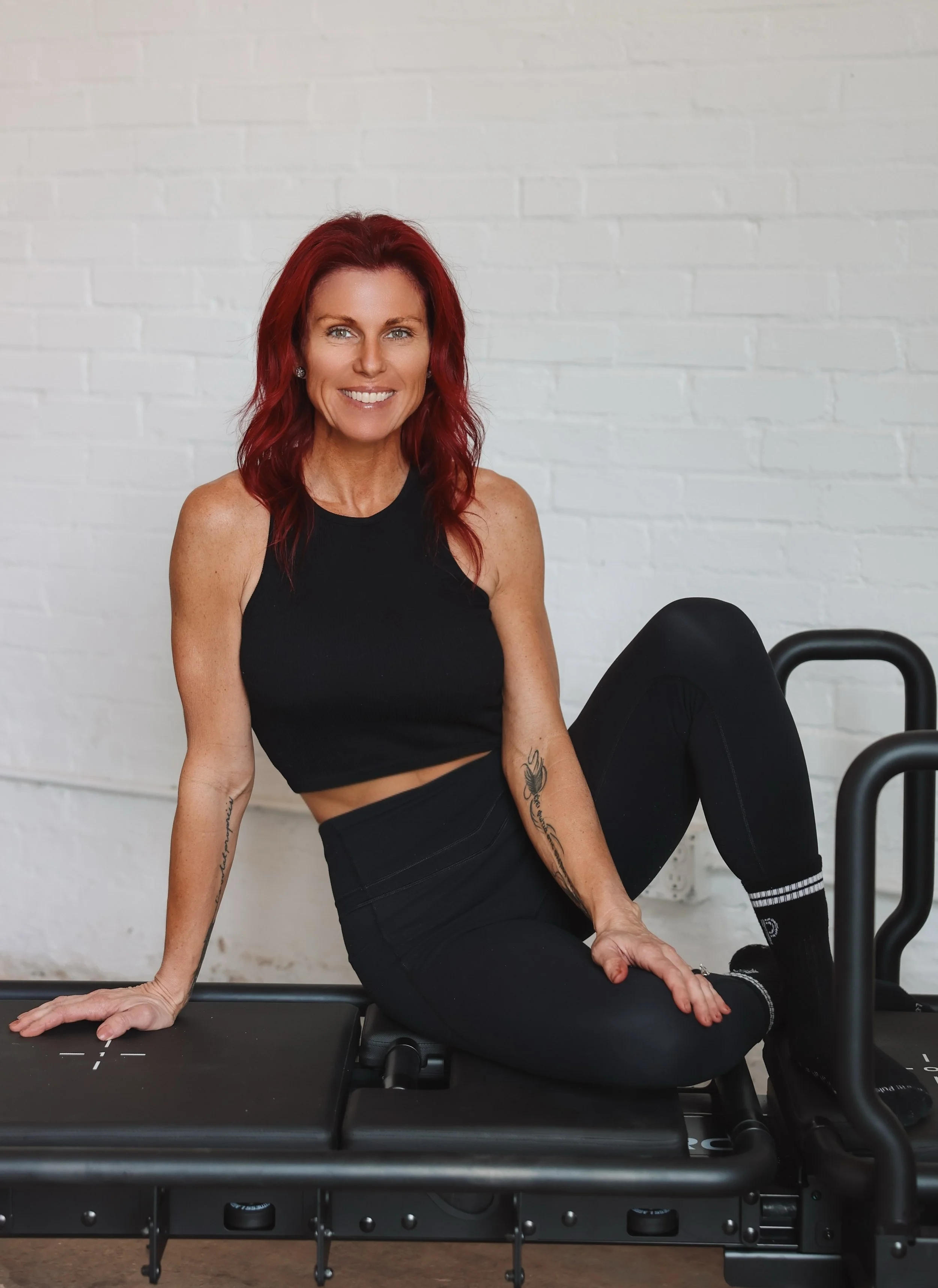 Karen Anaya is wearing a black athletic tank top and black leggings, is sitting on a Lagree Megaformer machine with a white brick wall in the background.