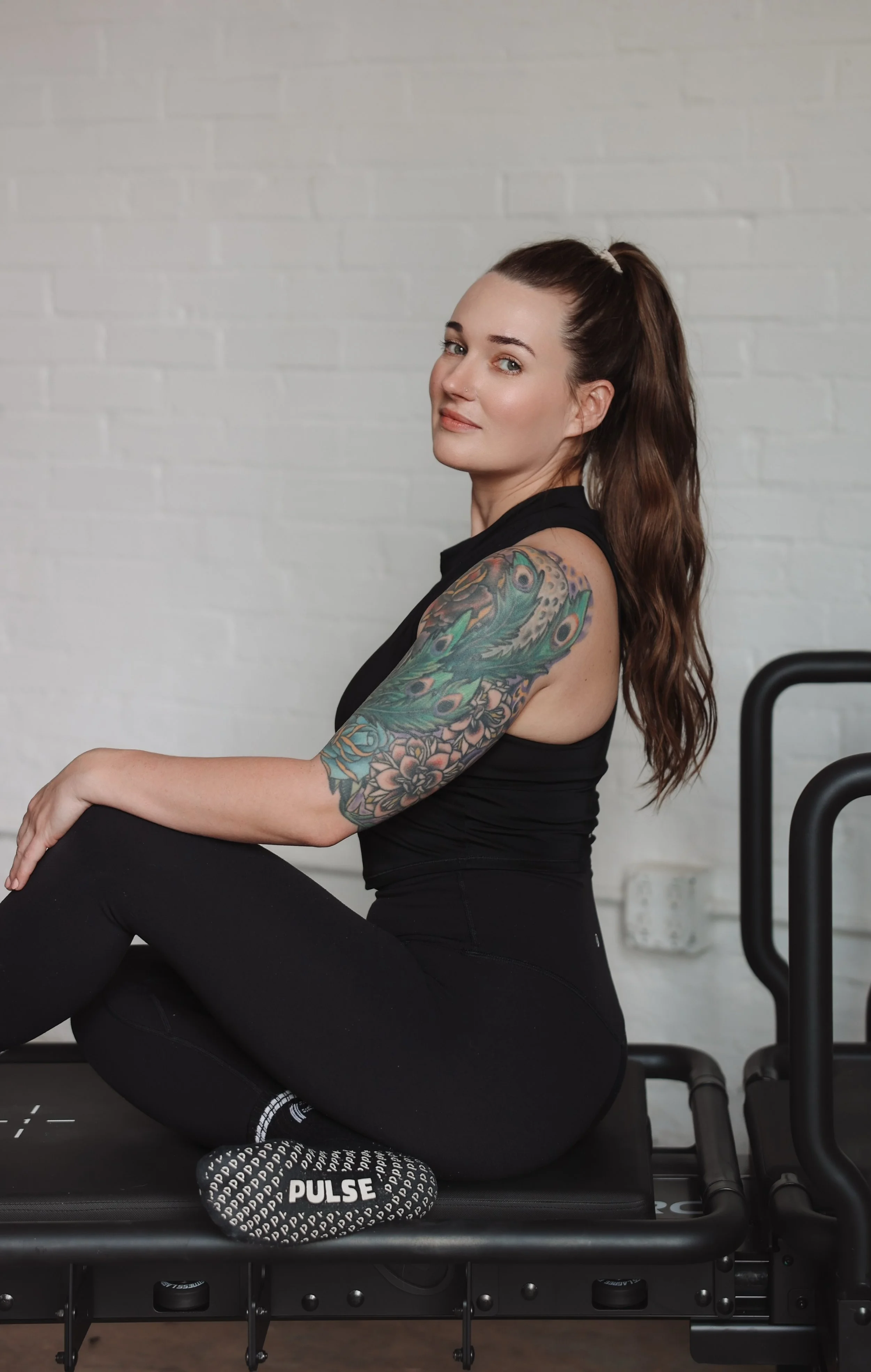 Shea Carver. Director of Pulse Lagree. She is sitting on a Megaformer against a white brick wall, wearing black athletic clothing and black and white Pulse branded grip socks.