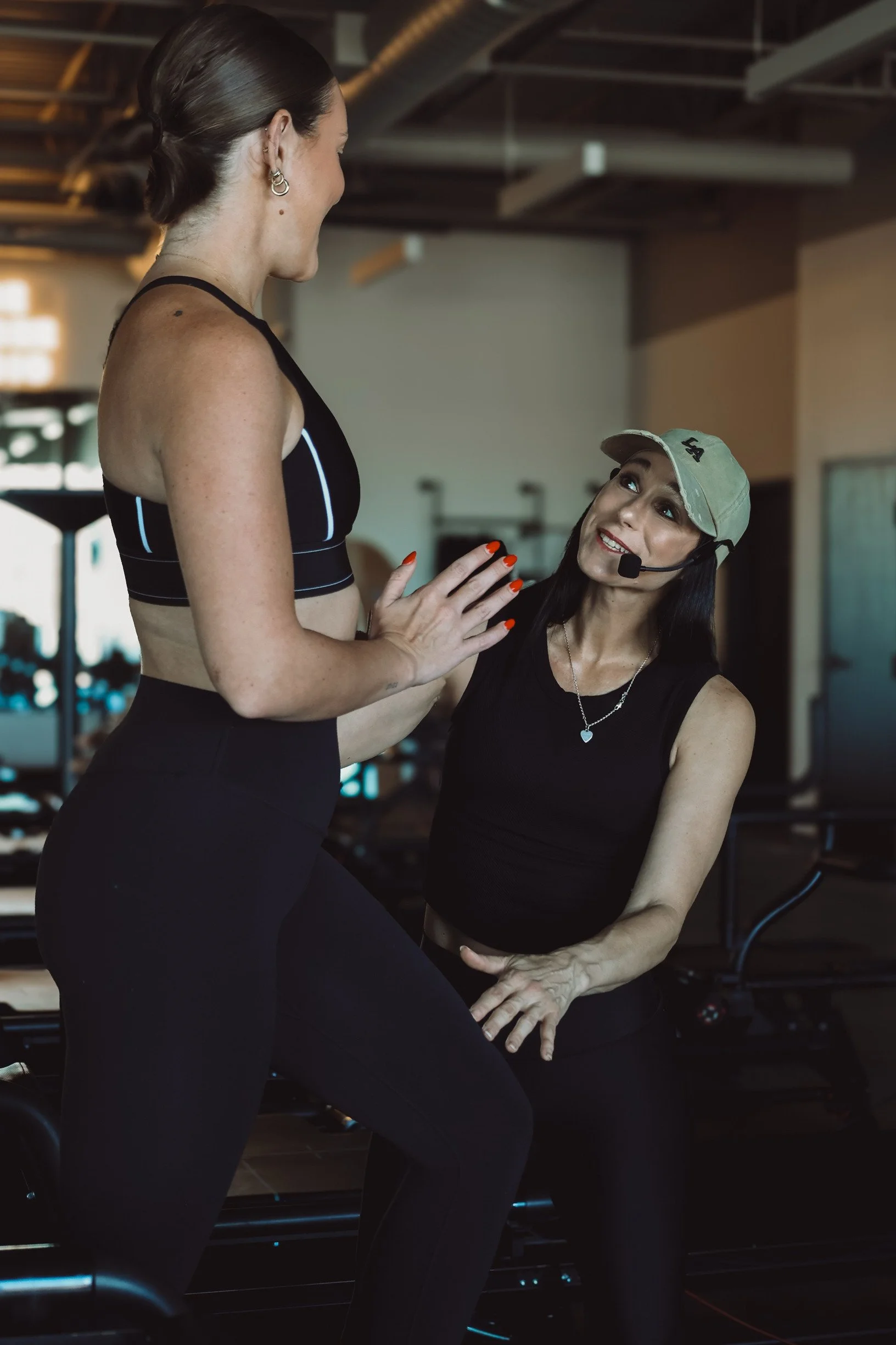 A personal trainer talking to a woman in a gym, both wearing athletic clothing, with the trainer holding a headset mic and the woman sitting on a piece of gym equipment.