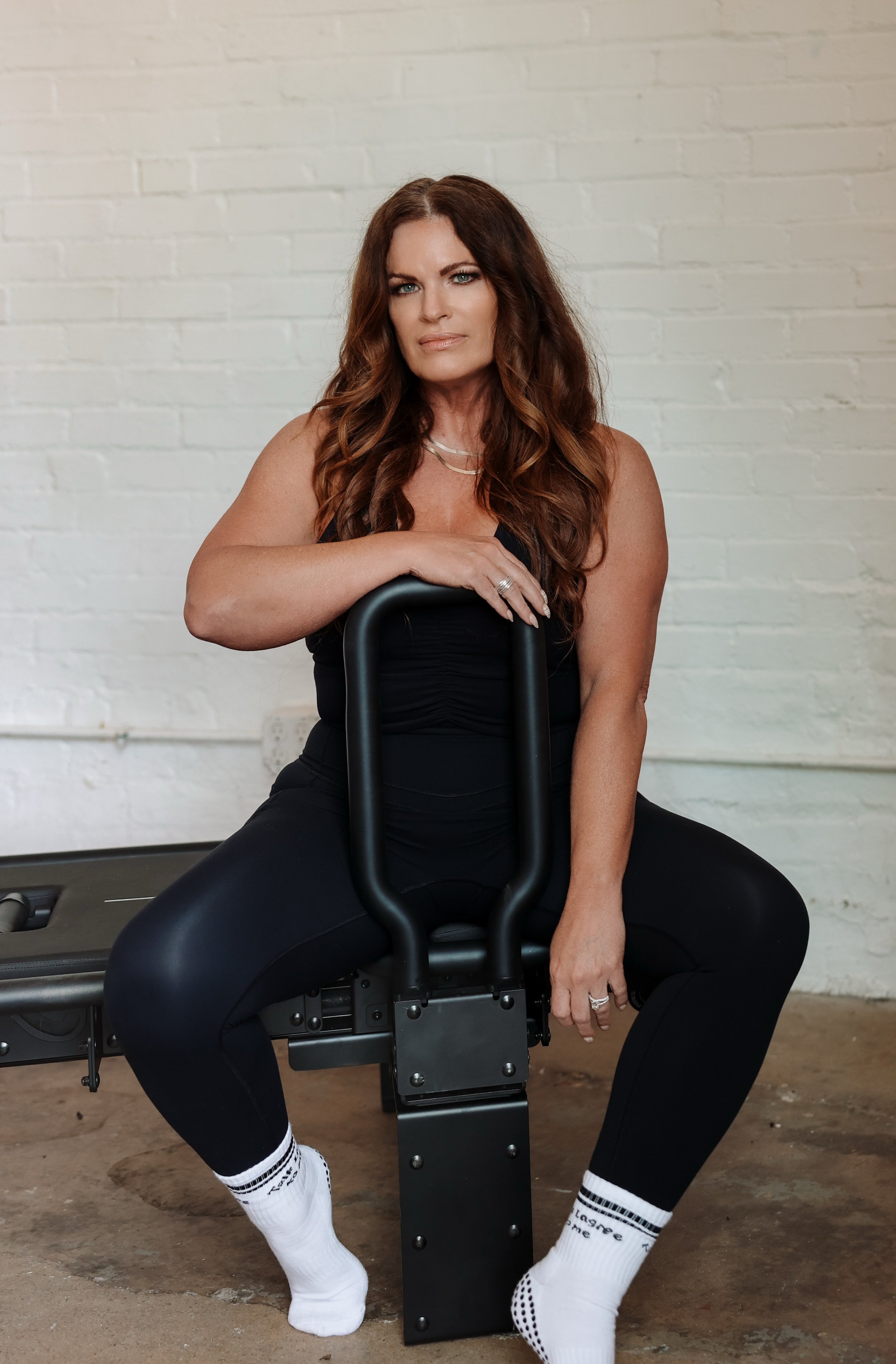 Stacy Praytor is wearing a black tank top and black pants, sitting cross-legged on a Lagree Megaformer machine in front of a white brick wall, wearing black athletic clothing.