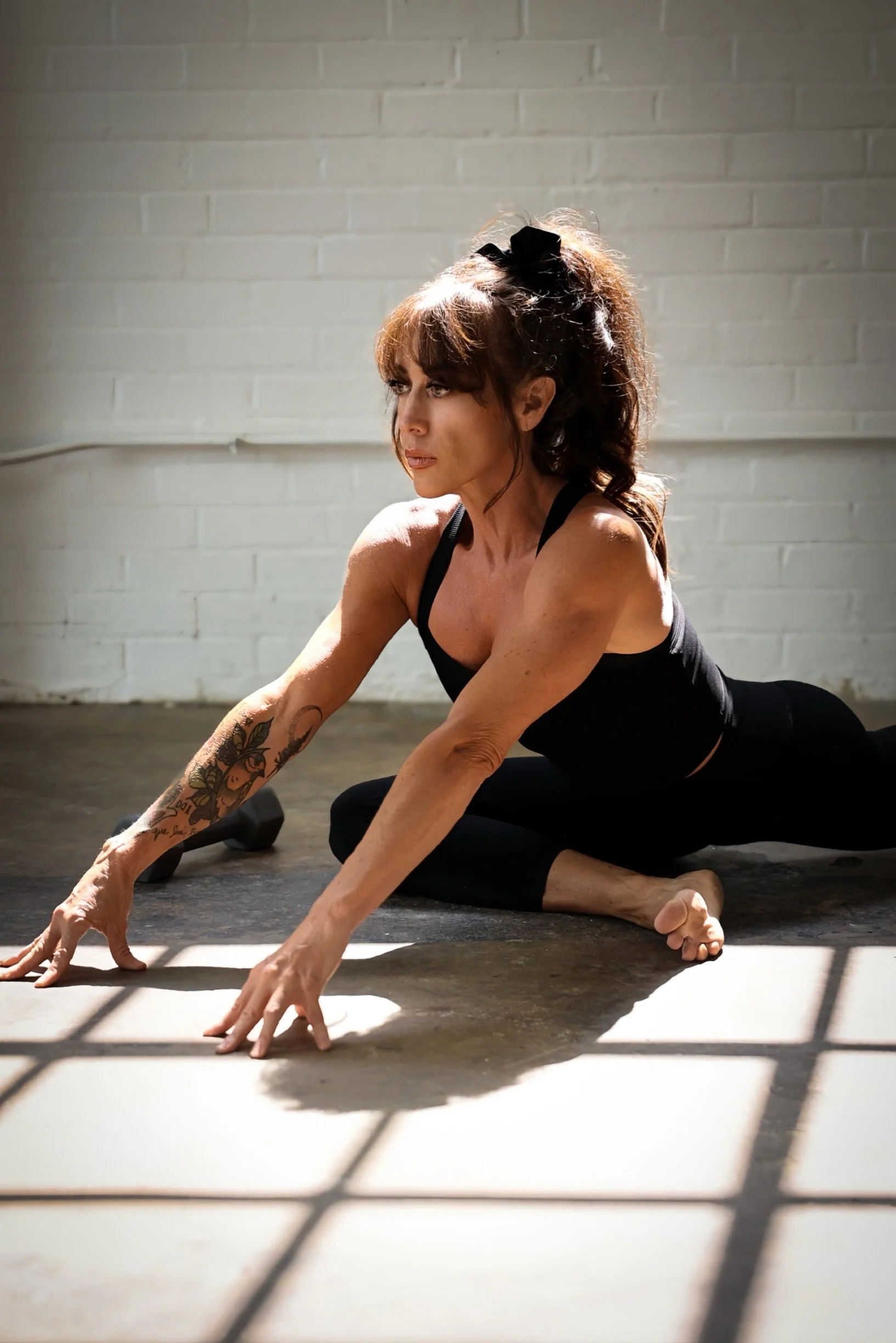 Shaena Campbell. A woman with tattoos on her arm practicing yoga on a black mat in a room with a white brick wall, sunlight casting shadows.