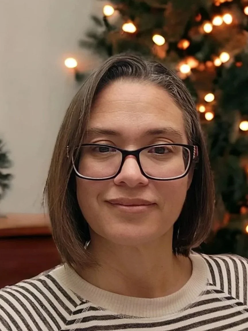 A woman with short brown hair and glasses smiling in front of a Christmas tree decorated with lights.
