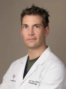 Portrait of a male doctor wearing a white lab coat against a neutral background.