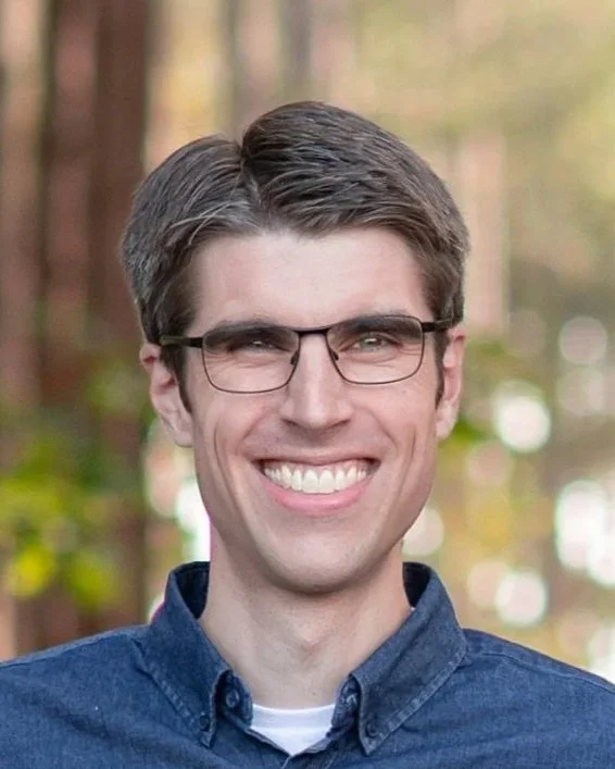 A young man with glasses smiling outdoors with blurred forest background.