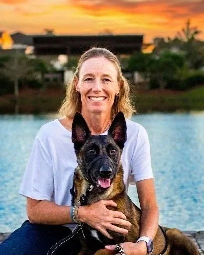 A smiling woman with blonde hair sitting outdoors at sunset, holding a Belgian Malinois dog in front of a body of water.
