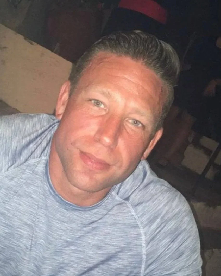 A smiling man with short, dark styled hair wearing a light grey athletic shirt, sitting indoors with a dark background.
