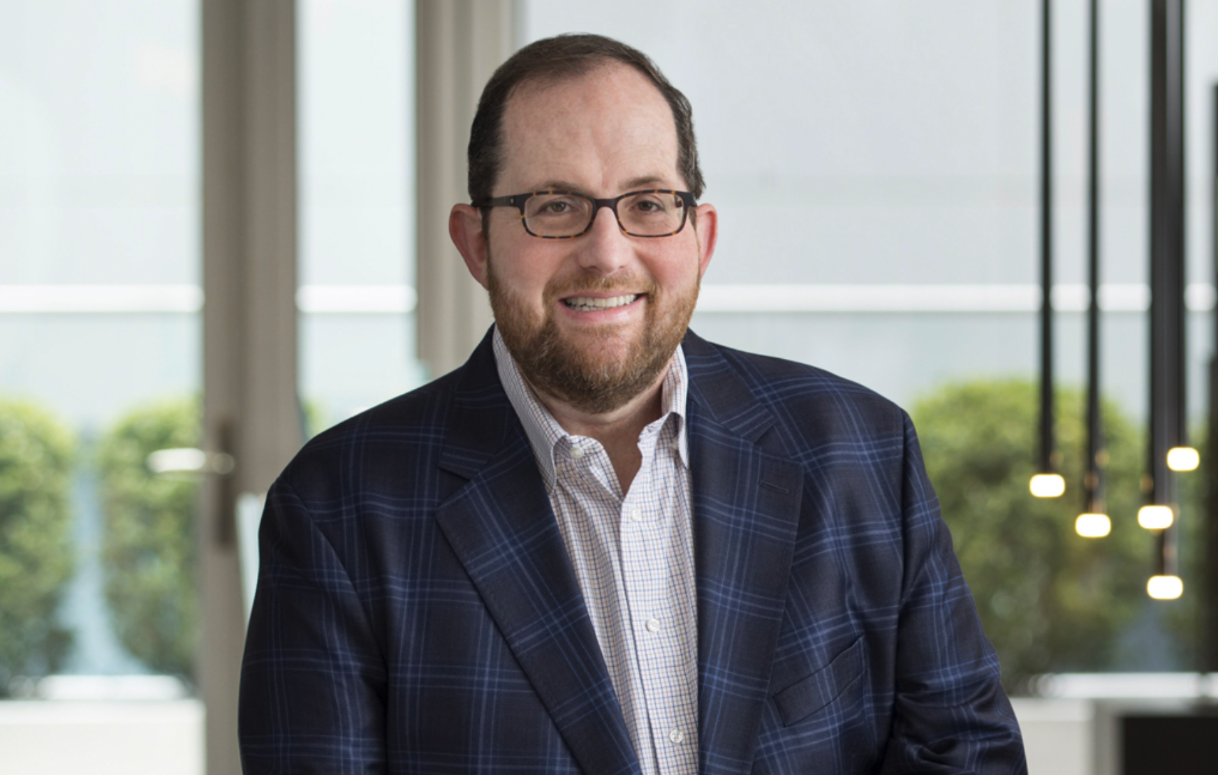 A man with glasses, a beard, and a smile, wearing a dark blue plaid suit and a light-colored button-up shirt, standing in a modern office setting with large windows and outdoor greenery visible in the background.