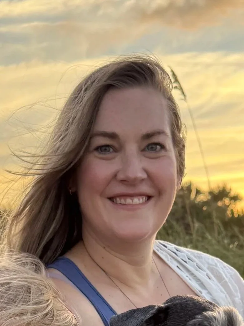 A woman with long, light brown hair smiling outdoors at sunset, wearing a blue top and white cardigan, with a small black dog partially visible in the foreground.