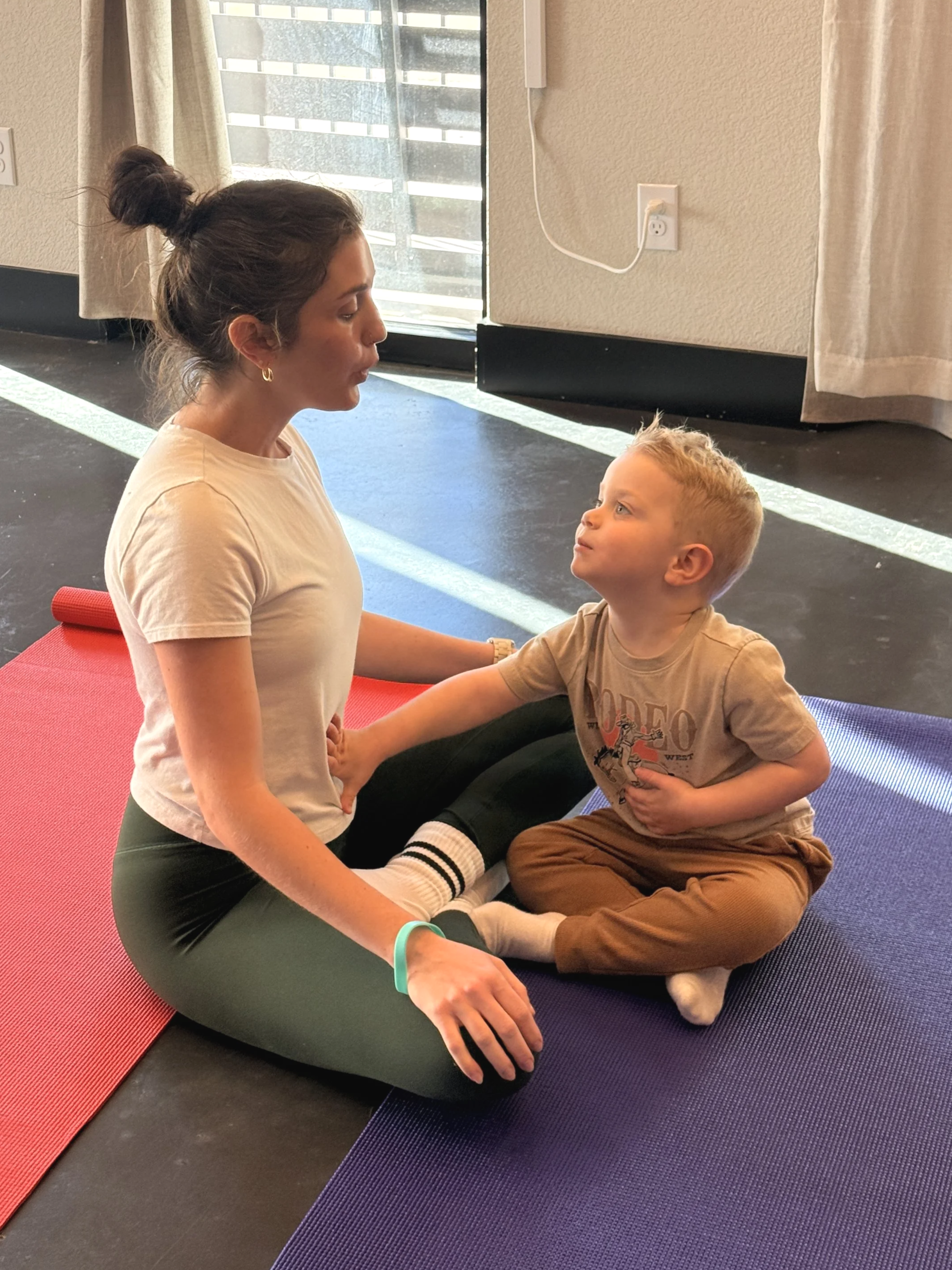 A woman and a young girl practicing yoga together in a bright, airy room with large windows, using yoga mats.