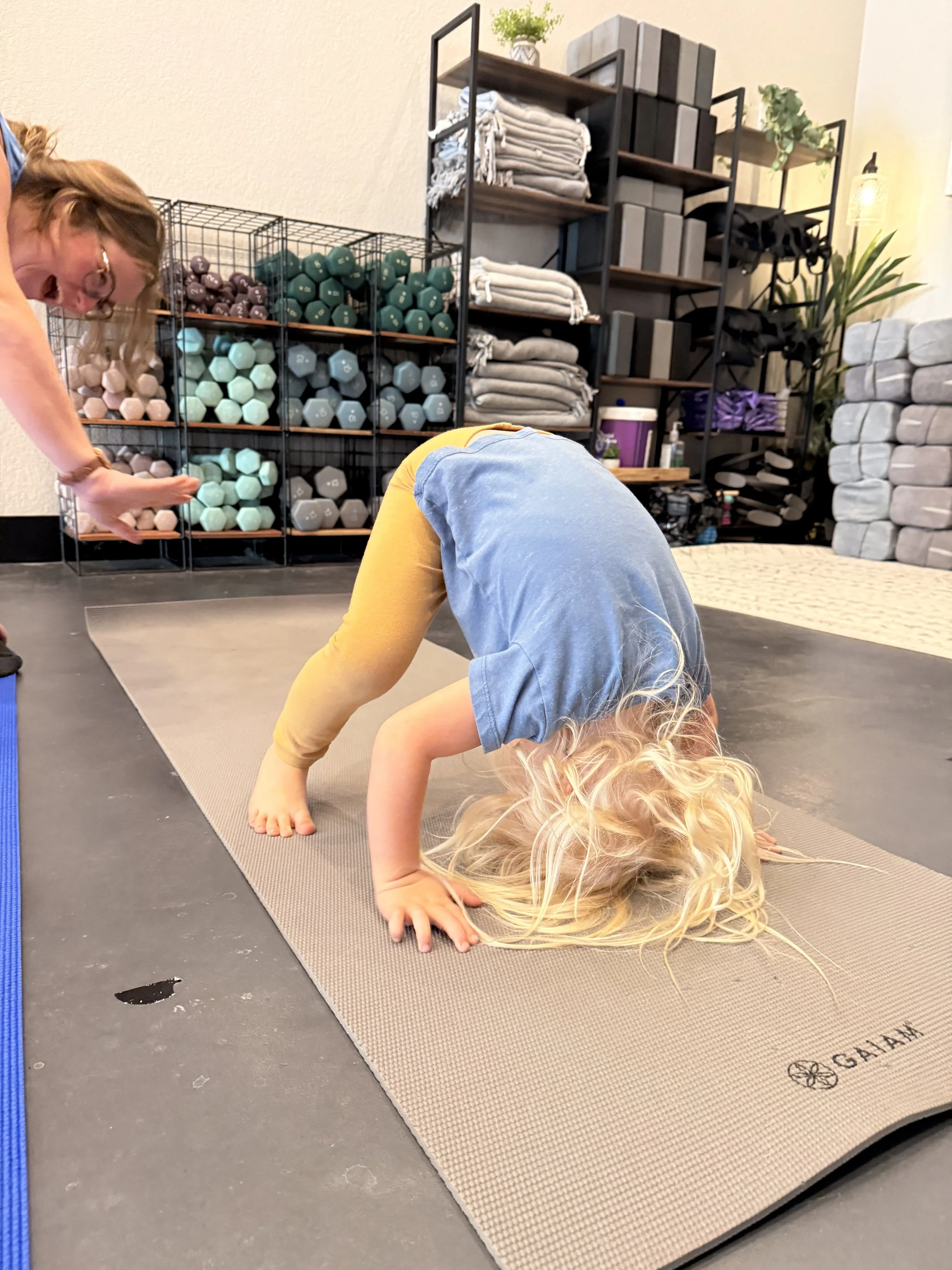 A woman and a young girl practicing yoga together in a bright, airy room with large windows, using yoga mats.