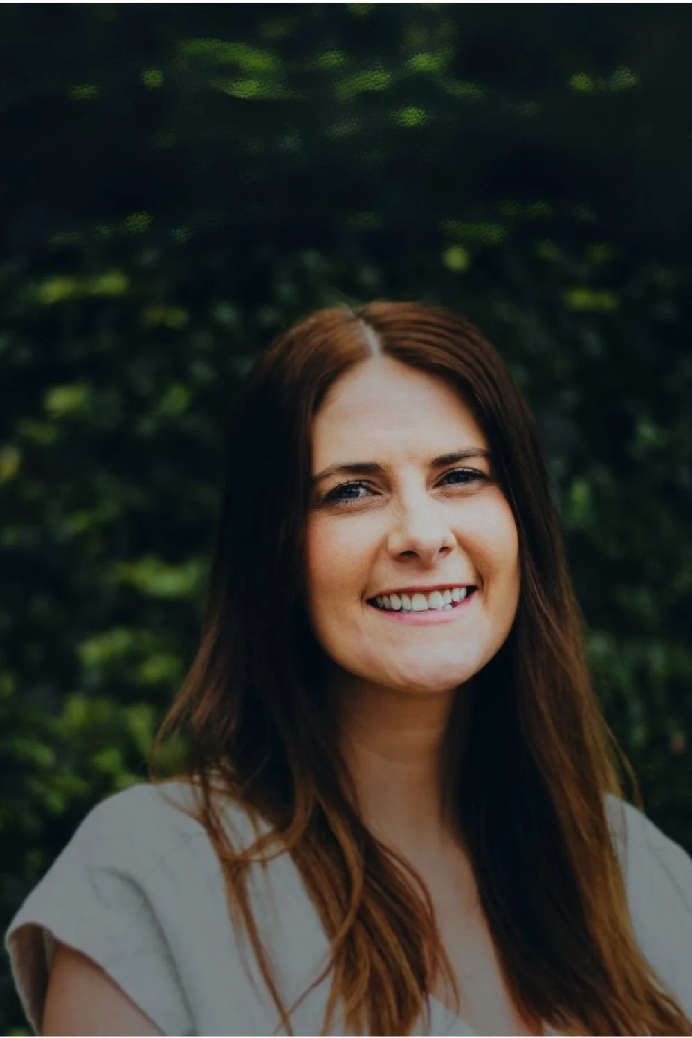 Close-up of a smiling woman with long, brown hair standing outdoors in front of green foliage.