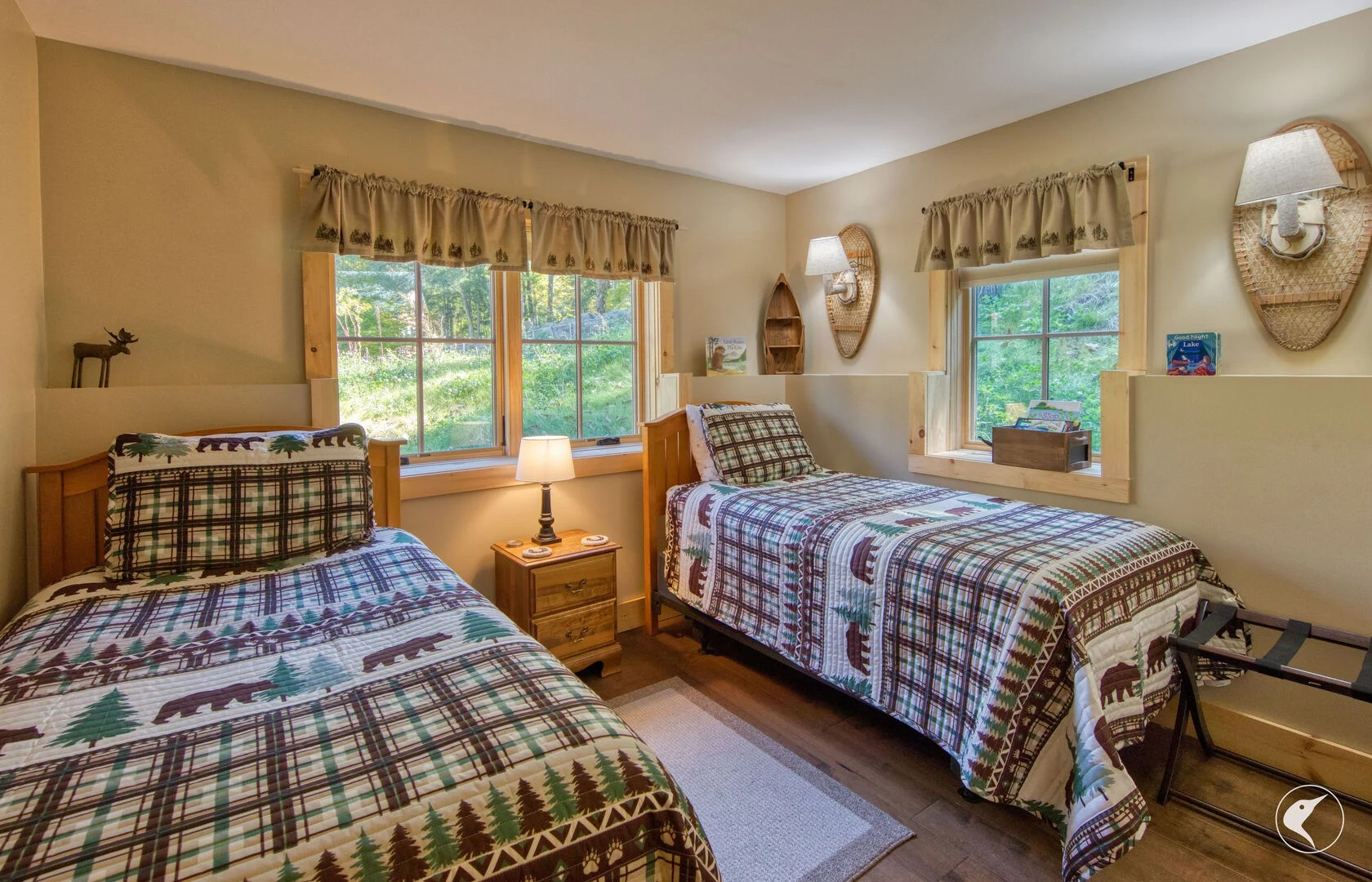 A cozy bedroom with two beds covered in plaid and moose-themed quilts, wooden furniture, and windows showing a green, forested outdoors.