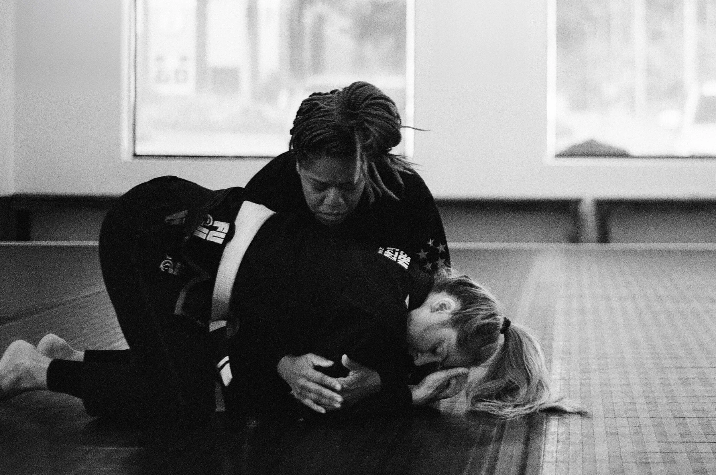 Two women practicing Brazilian Jiu-Jitsu on a mat in a training facility; one woman is executing a submission hold on the other, who is on her back with her head turned to the side.