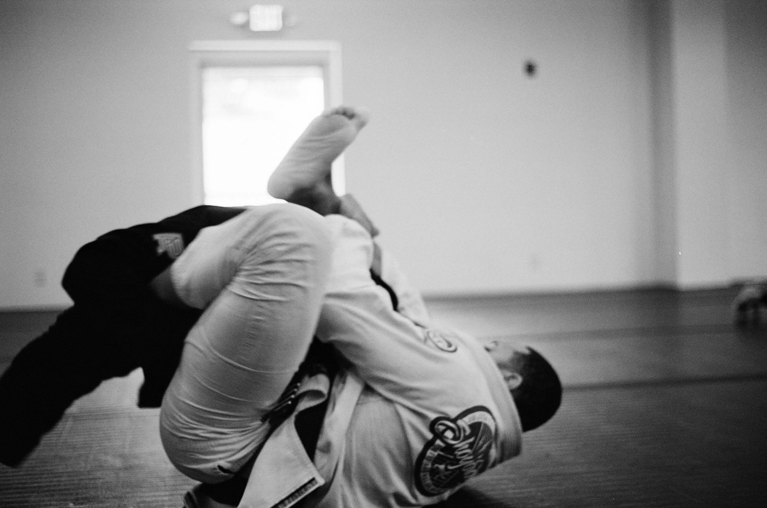 Two people practicing Brazilian jiu-jitsu on a mat, with one person on top and the other on the ground, in a training room.