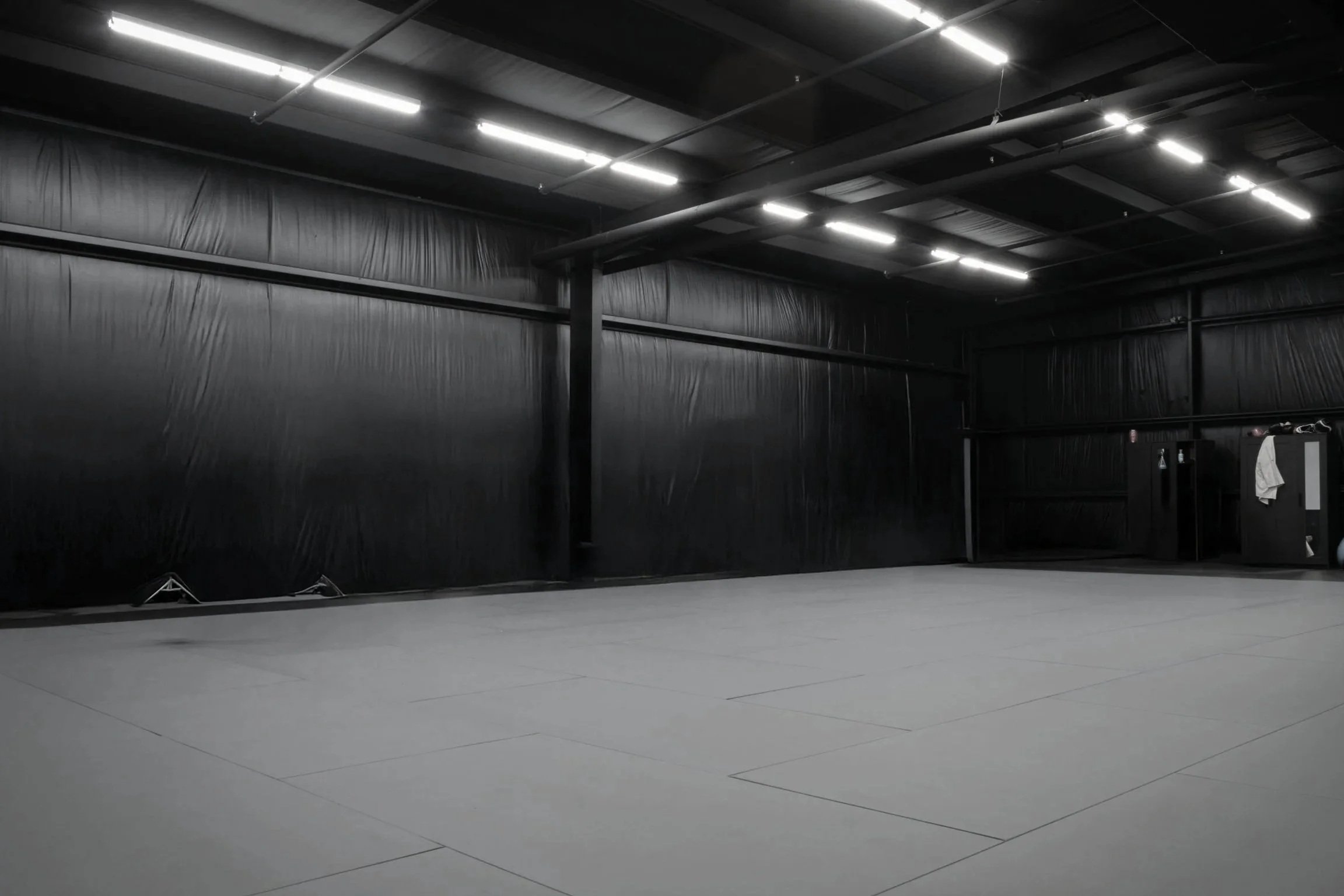 Empty black martial arts training gym with padded flooring and black walls, illuminated by fluorescent lights.