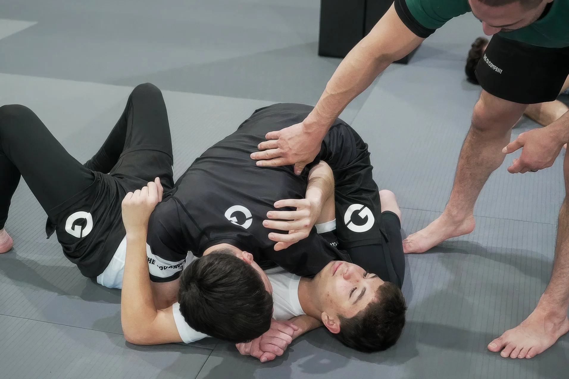 Two individuals practicing Brazilian Jiu-Jitsu on a gray mat, with one person on the ground in a black Gi and the other in a white Gi, surrounded by other participants and an instructor.