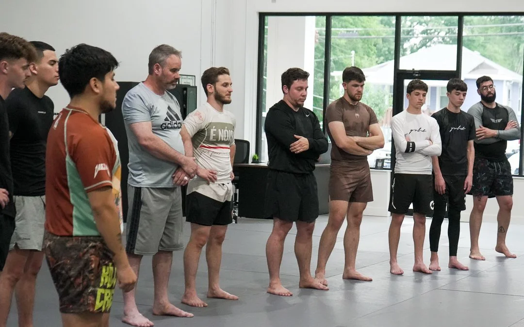 A group of nine men standing in a line on a martial arts mat inside a gym, facing forward with their arms crossed or at their sides, near large windows showing trees outside.