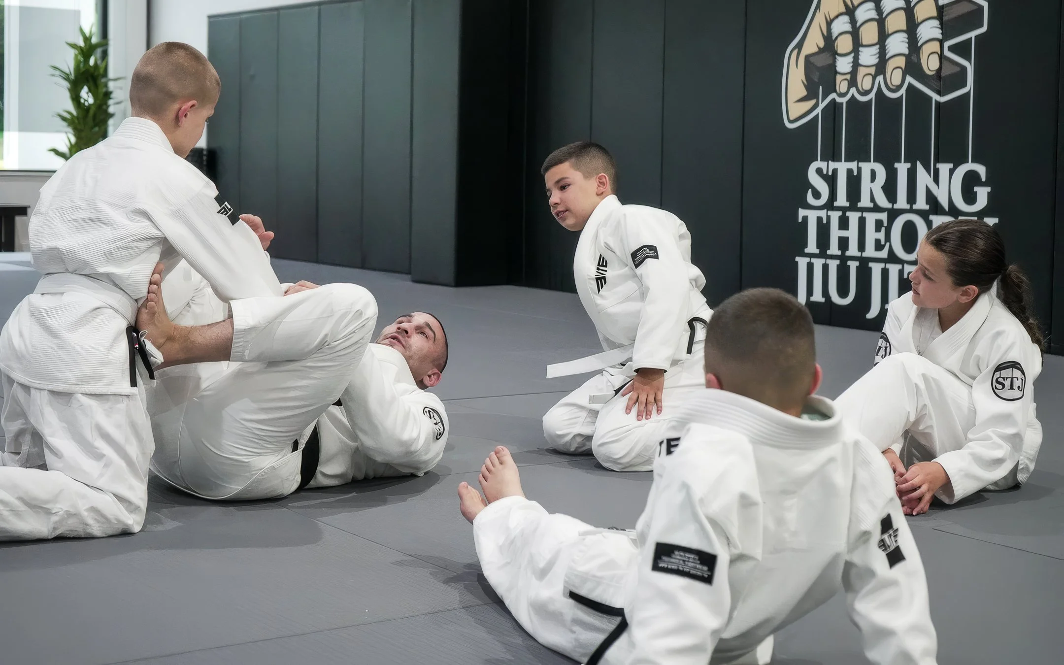 Children and an adult practicing Brazilian Jiu-Jitsu in a dojo, with a large black wall featuring the words "String Theory Jiu-Jitsu" and a graphic of a hand with fingers wrapped in tape.