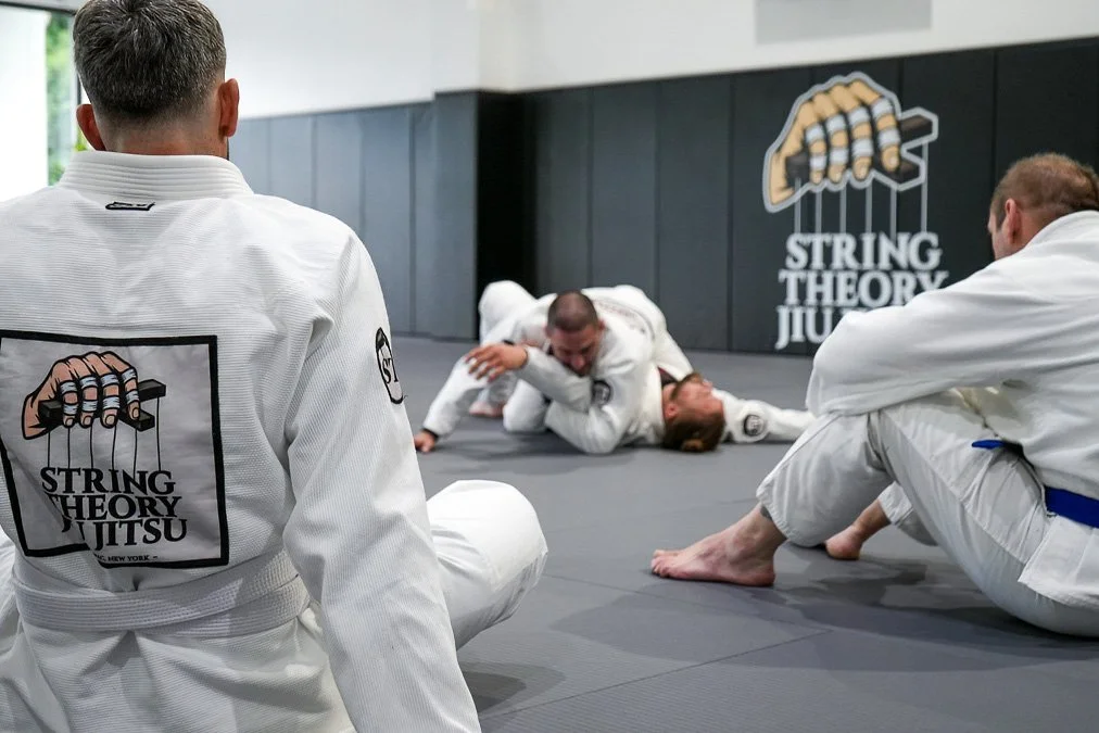 Two pairs of Brazilian Jiu-Jitsu practitioners are training on the mat, with one pair engaged in a grappling position in the background. In the foreground, two other practitioners sit on the mat observing. The logo on the gi reads 'String Theory Jiu Jitsu'.