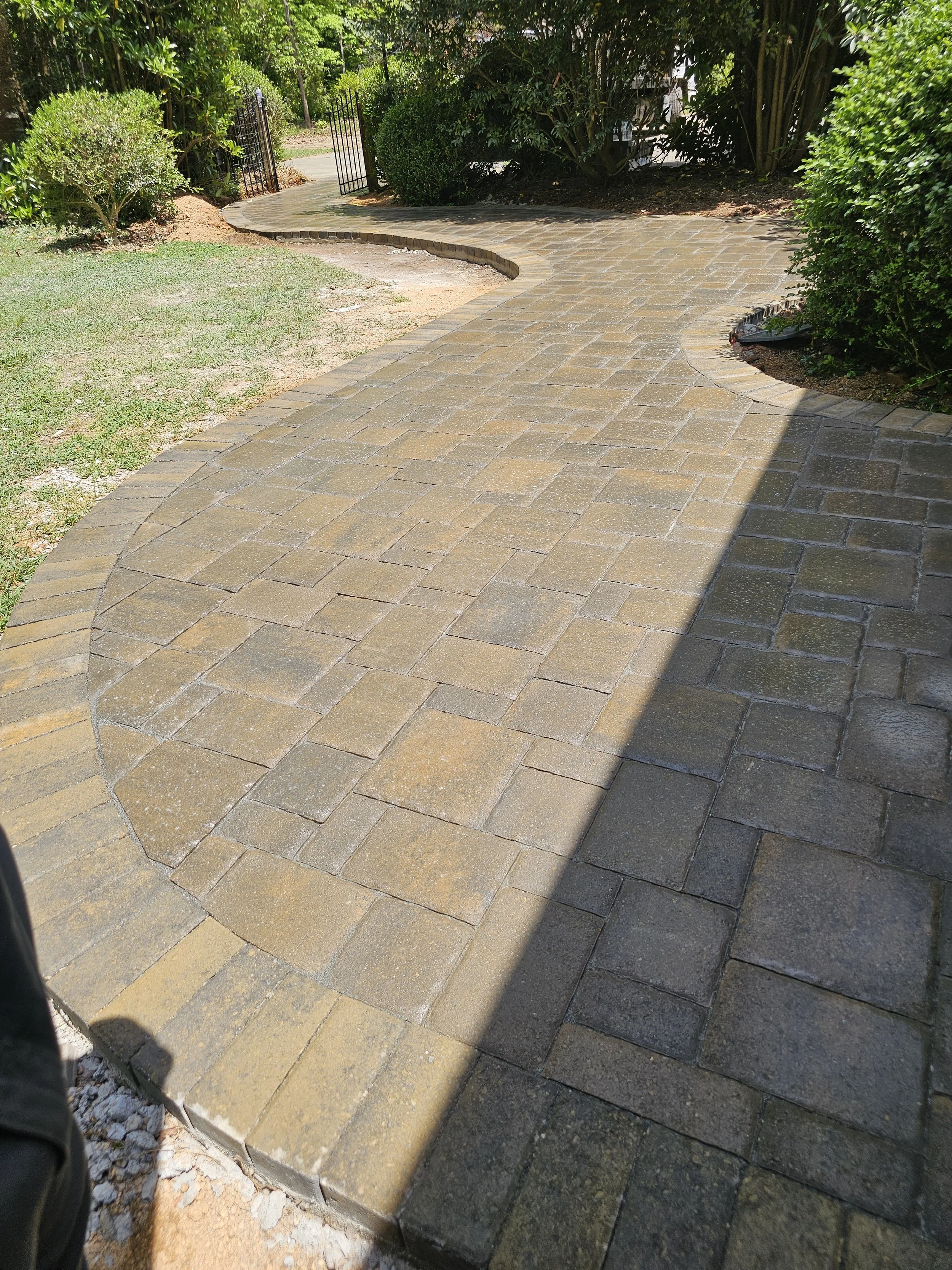 A curved brick walkway through a landscaped garden with green bushes and trees, and a black metal gate at the background.