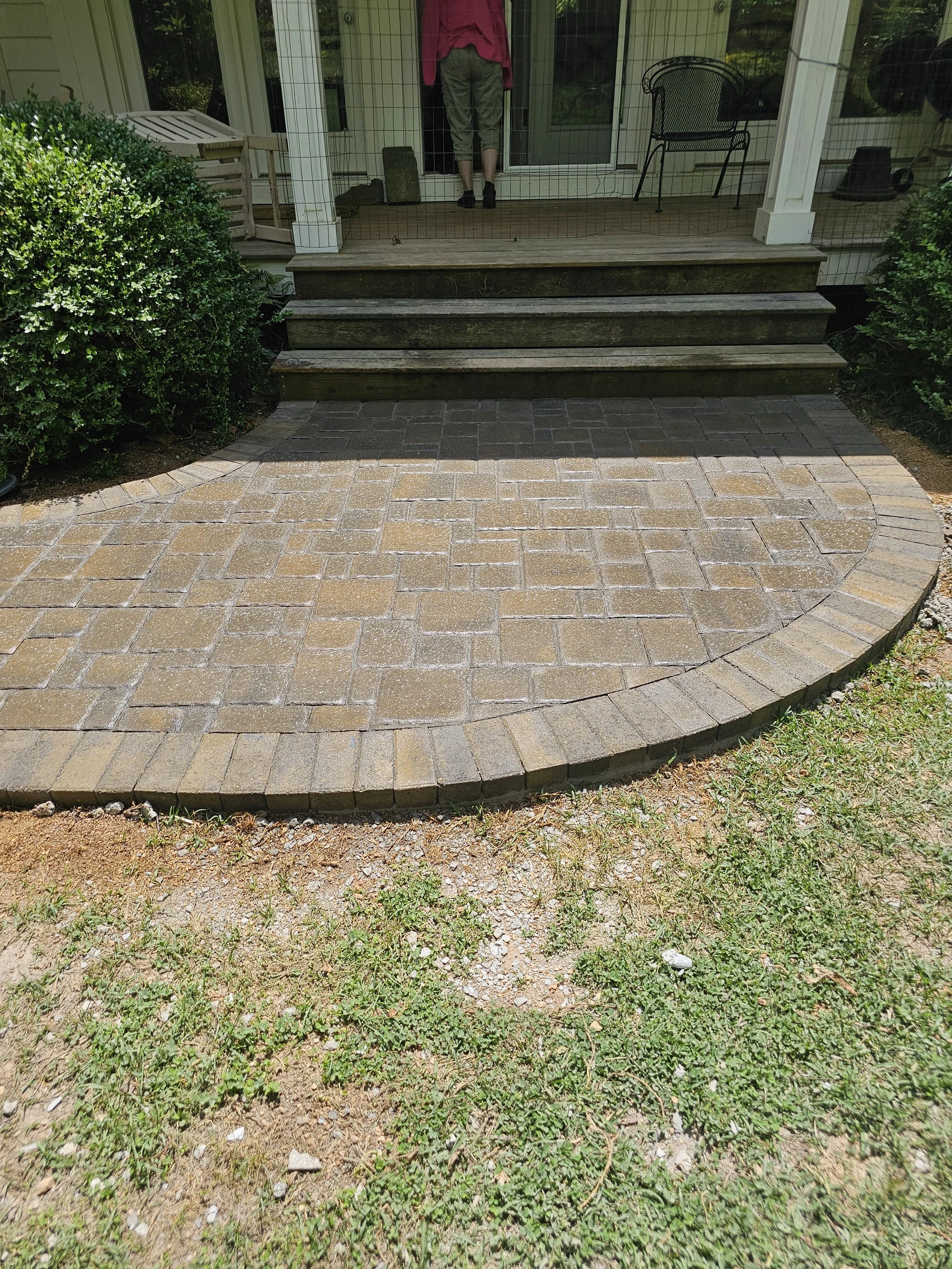 Newly installed brick patio in front of a house with steps, shrubs on both sides, and a person standing on the porch.