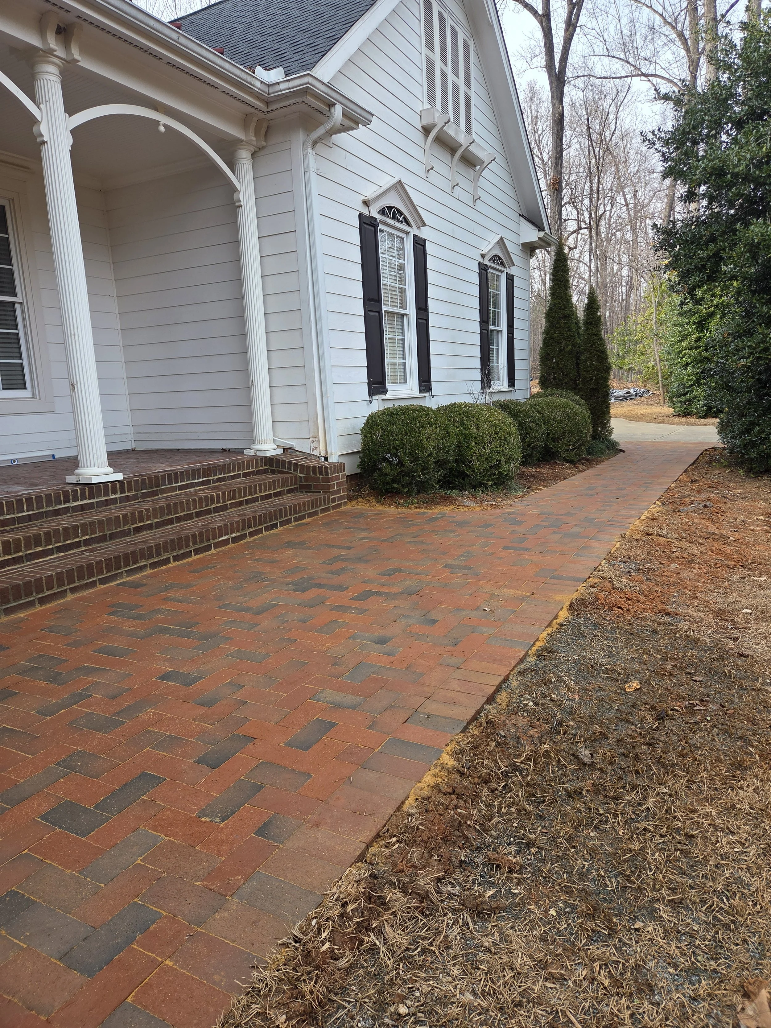 A brick pathway leading up to the front porch of a white house with black shutters, surrounded by manicured bushes and trees, on a slightly overcast day.