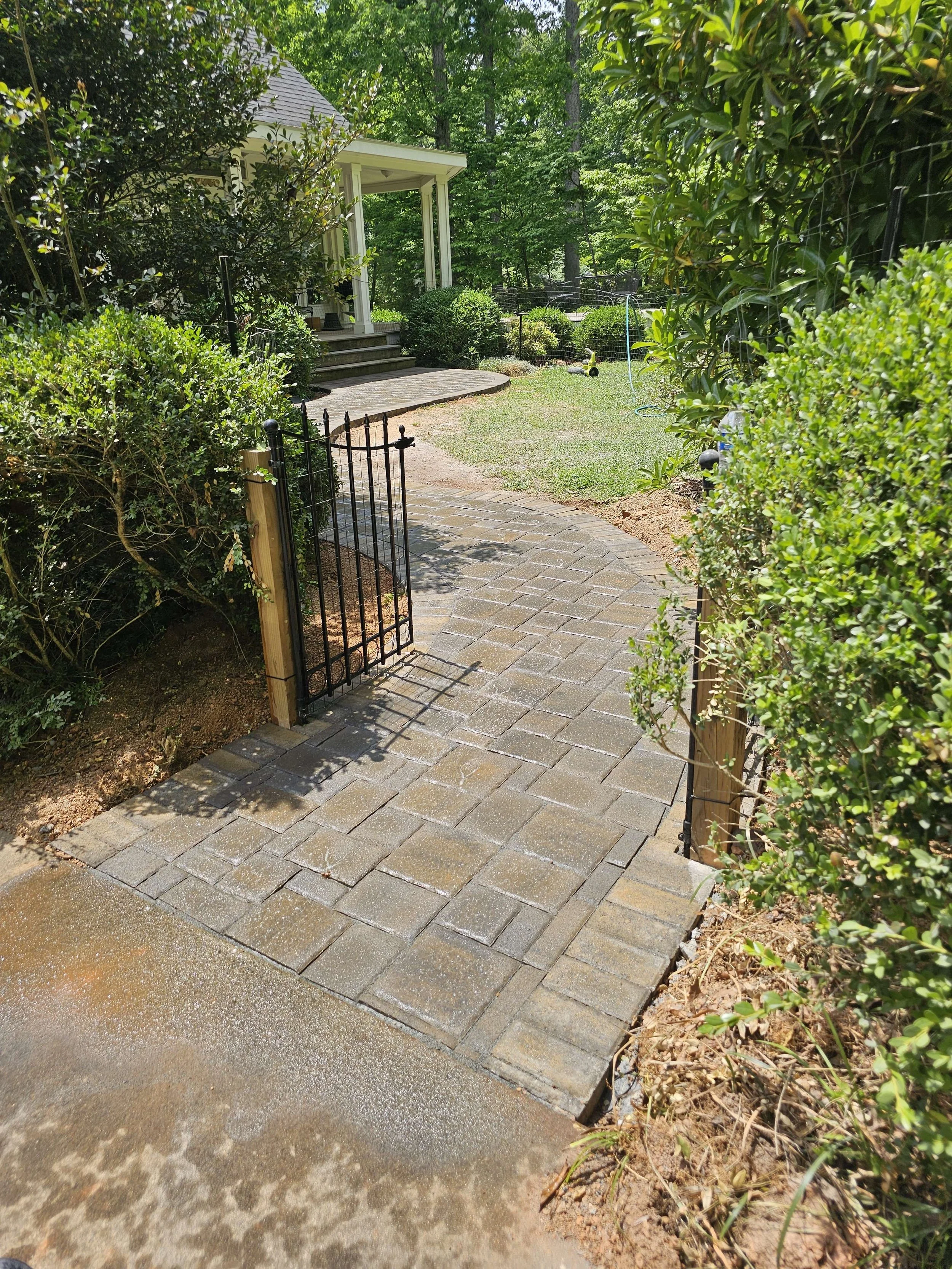 A brick pathway leading to a backyard yard with a small black metal gate, surrounded by bushes and trees, with a house porch visible in the background.