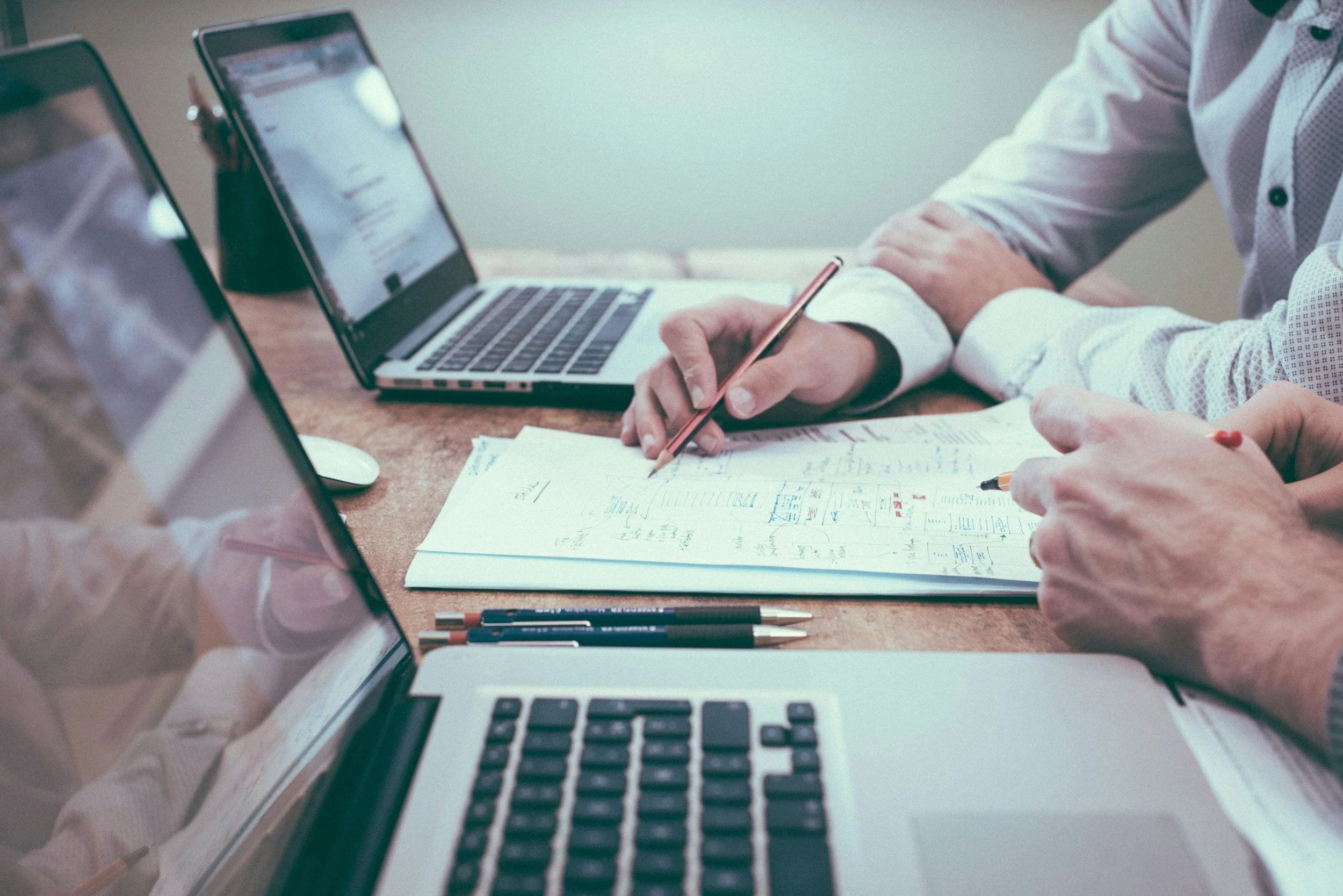 Two people working at a desk with laptops and printed documents, taking notes and discussing.